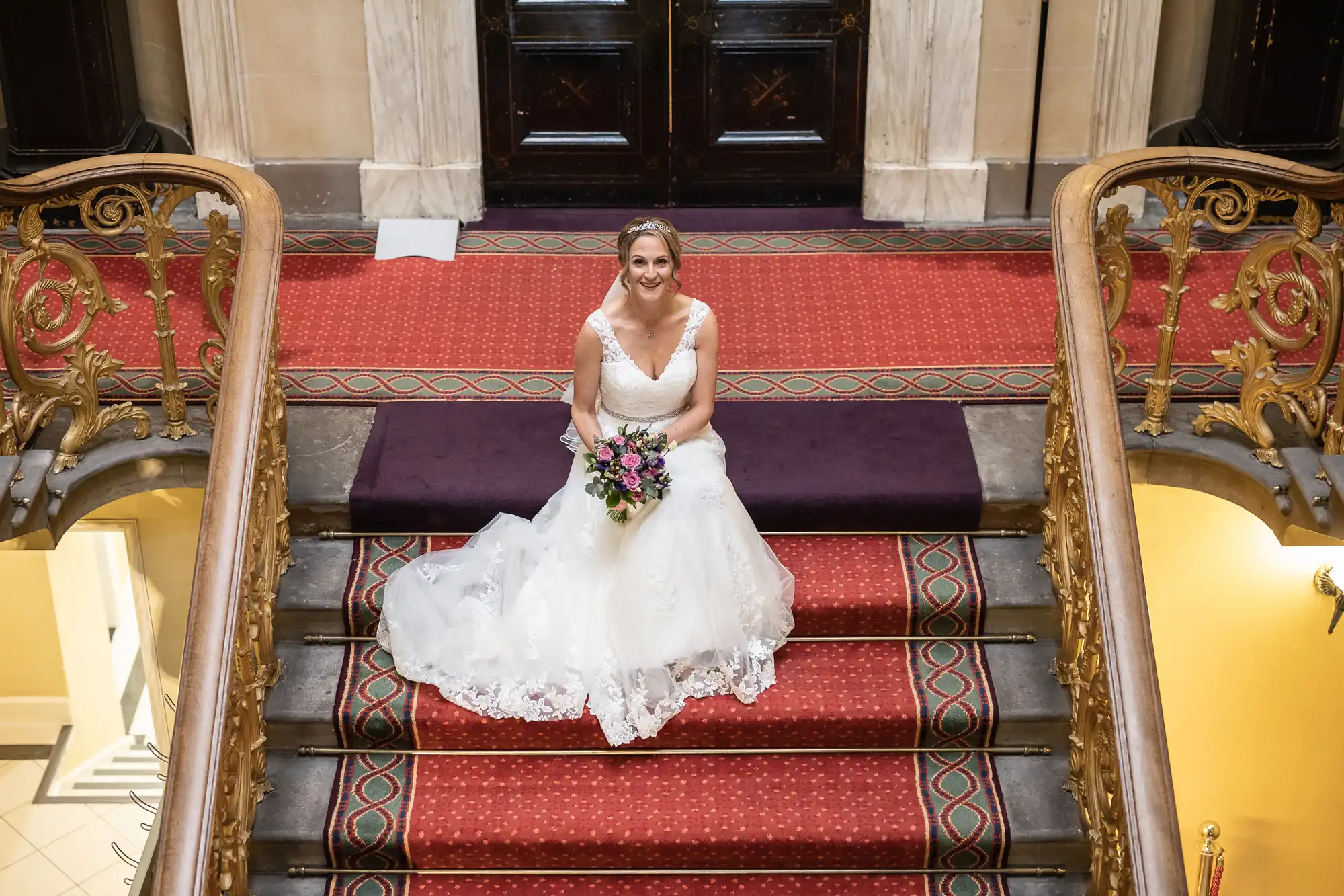 A woman in a white wedding dress sits on the steps of a grand staircase with red carpeting, holding a bouquet of flowers and smiling.