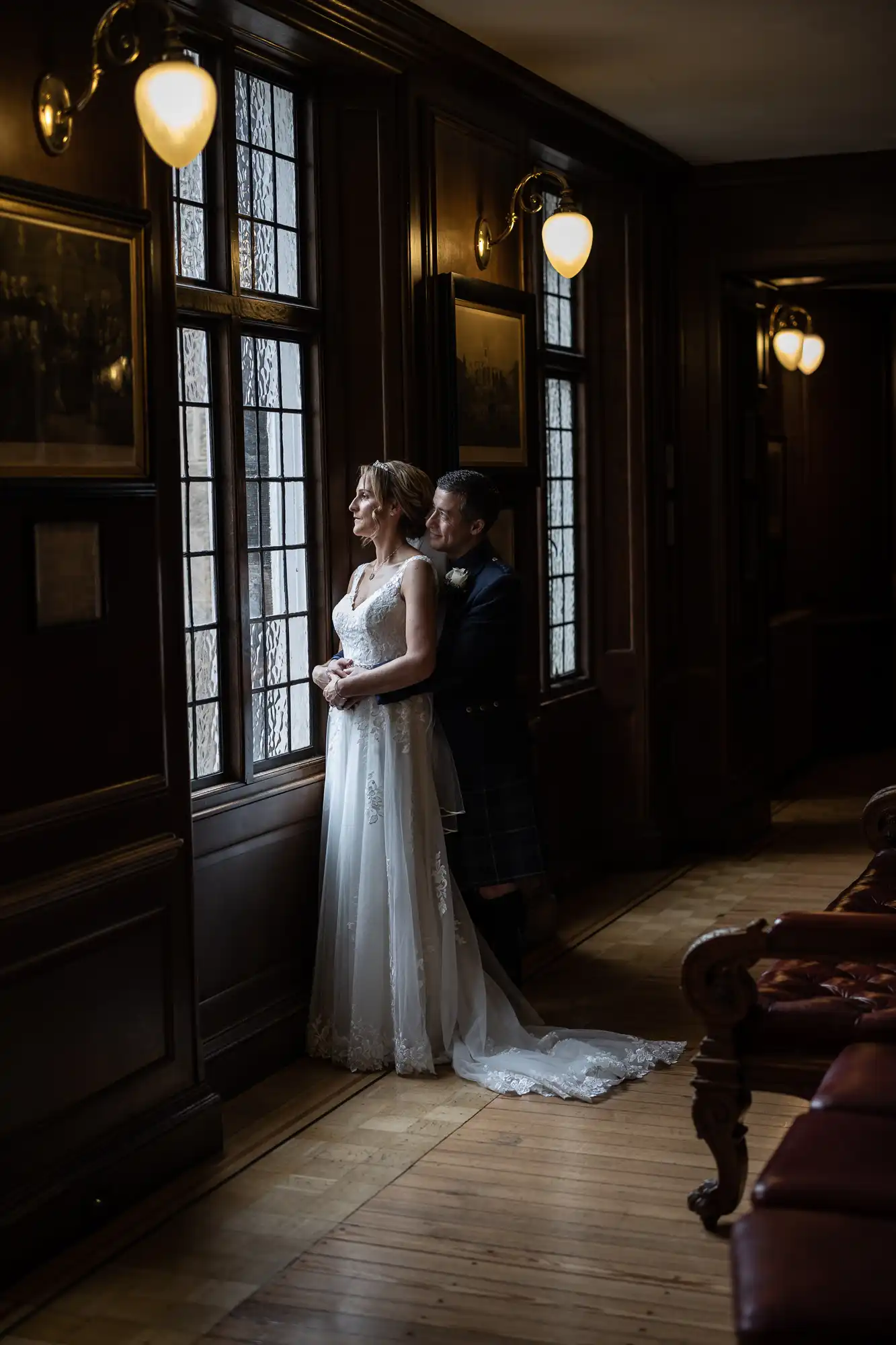 A bride and groom stand together by a window in a dimly lit room with wooden walls and framed pictures.