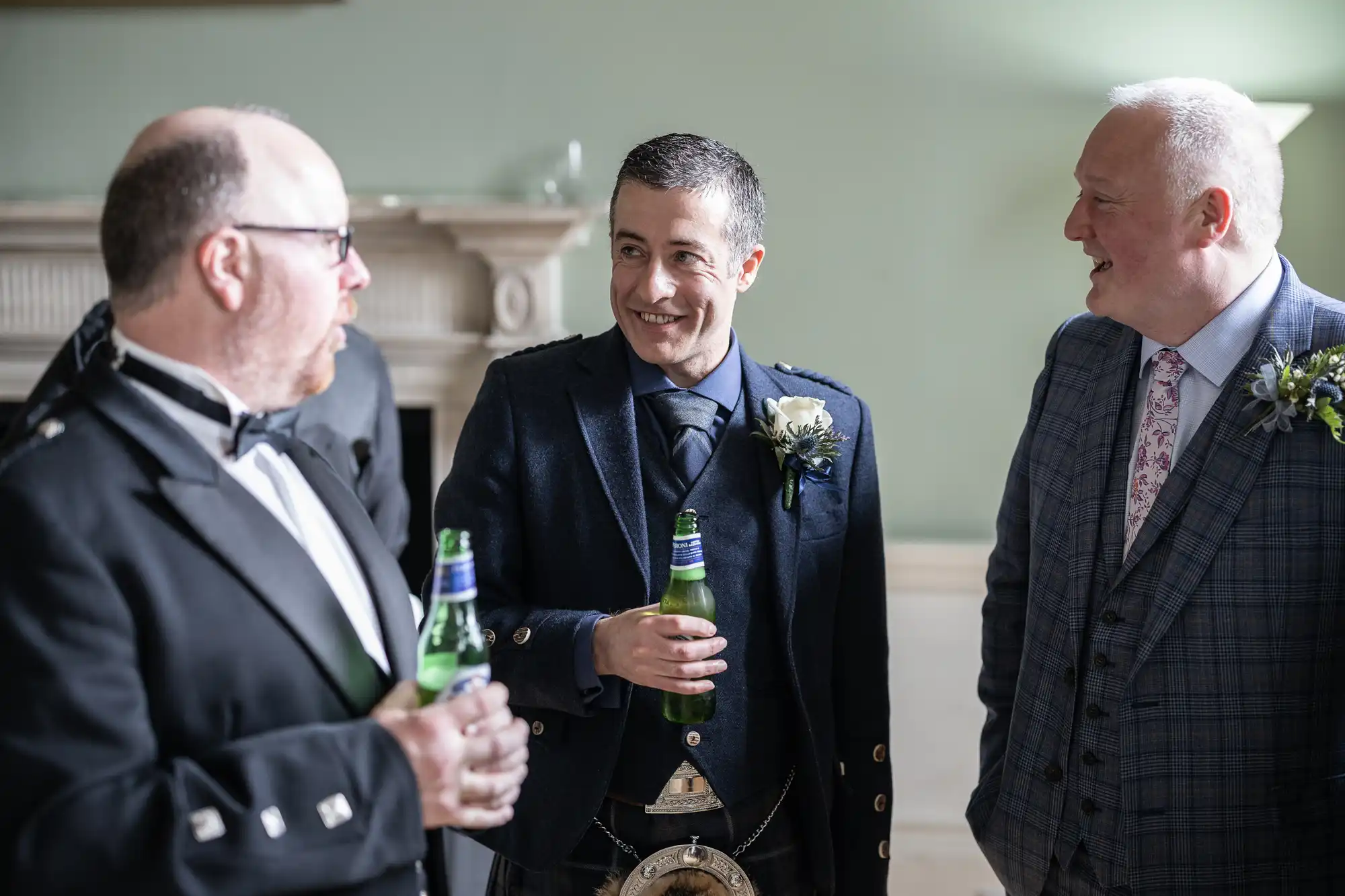 Three men in suits stand indoors, holding beer bottles and chatting. One man, in the middle, wears a traditional Scottish outfit with a sporran.