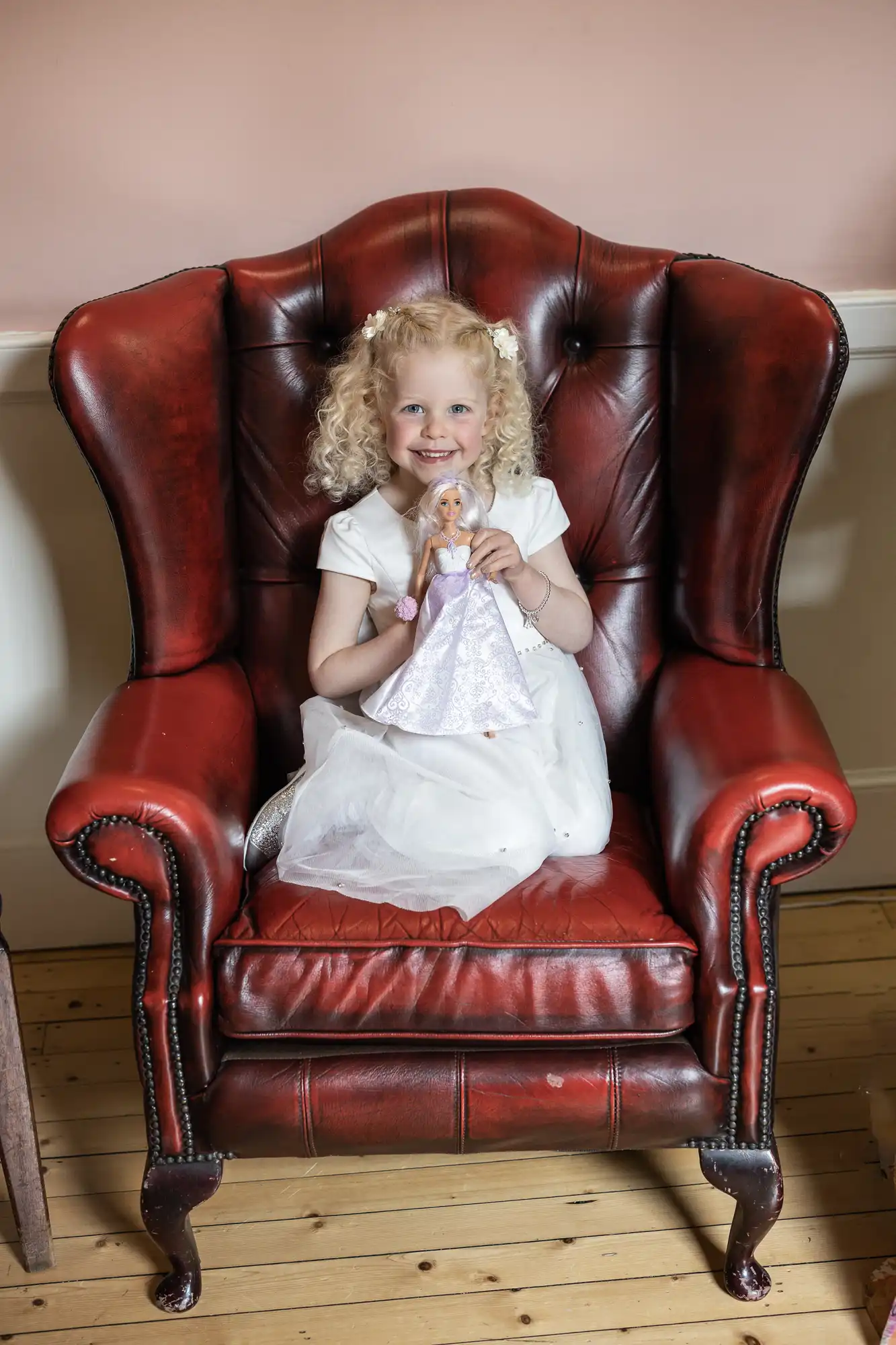 A young girl with curly blond hair sits on a large red leather armchair, holding a doll and smiling at the camera. She is wearing a white dress.