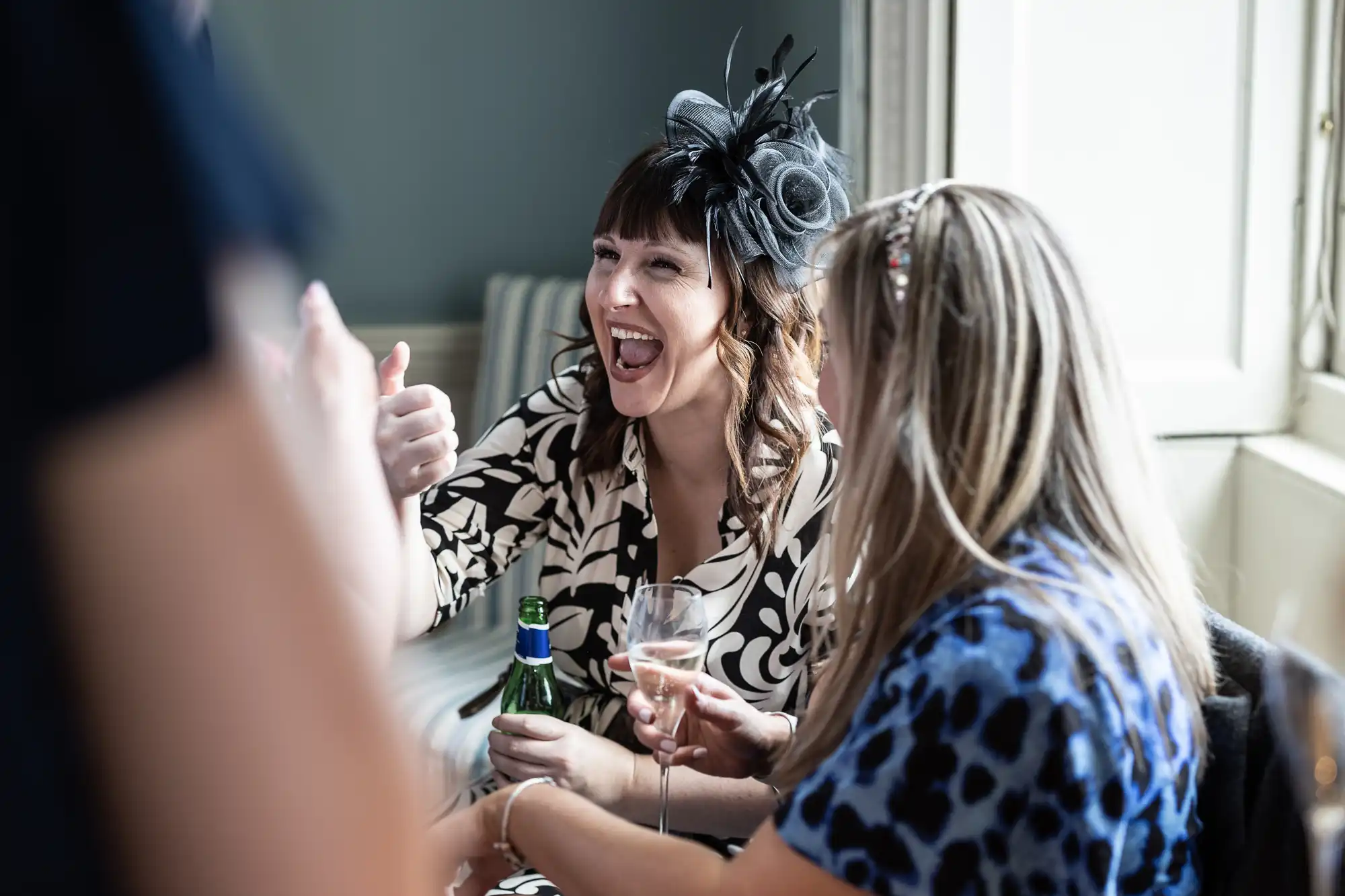 Two women conversing at a social event; one wearing a patterned dress and fascinator, giving a thumbs-up while laughing, the other woman holding a drink and wearing a blue patterned dress.
