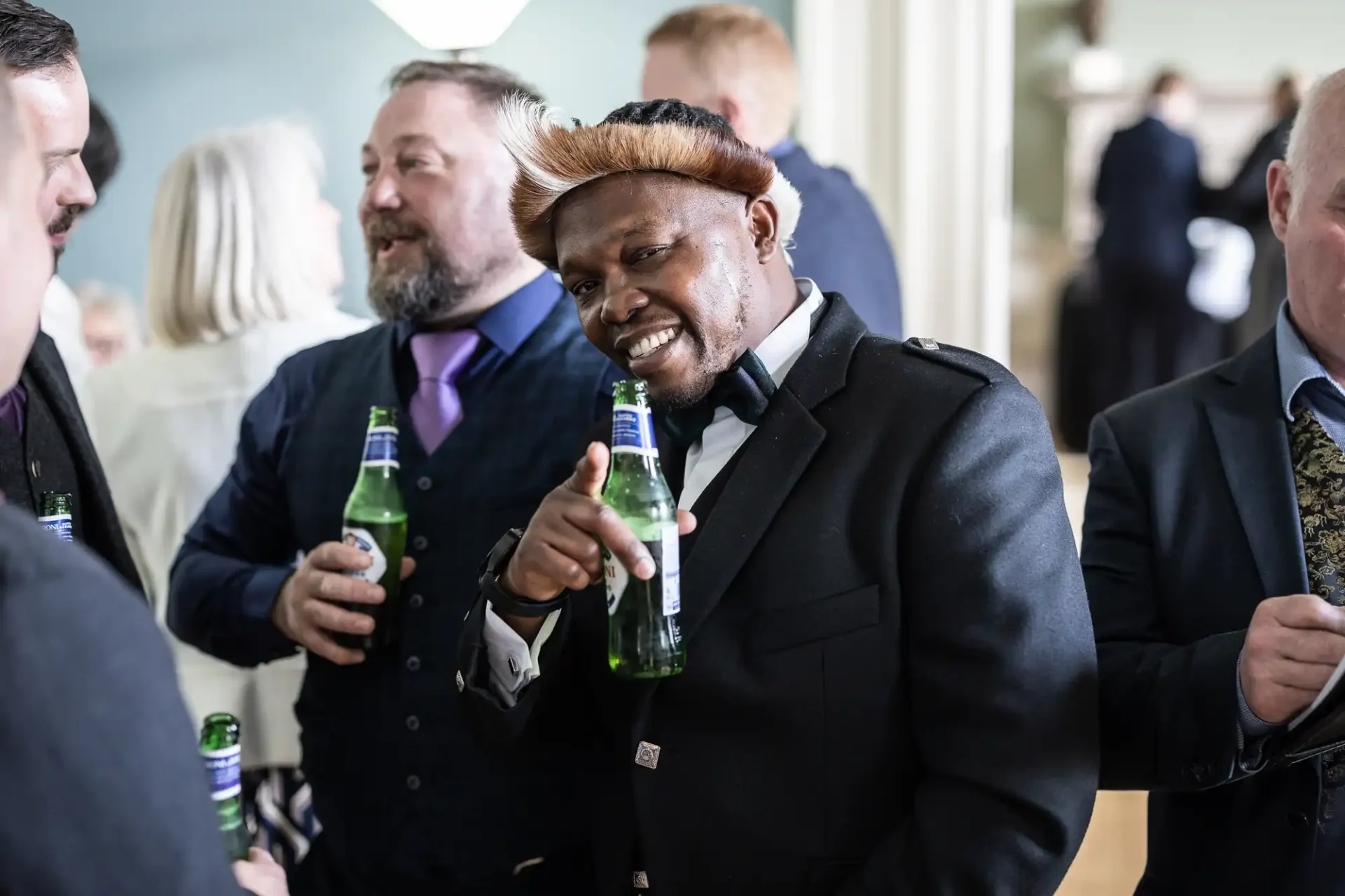 Several people socialize, holding bottled drinks. A man in a black suit and traditional hat points towards the camera while smiling.