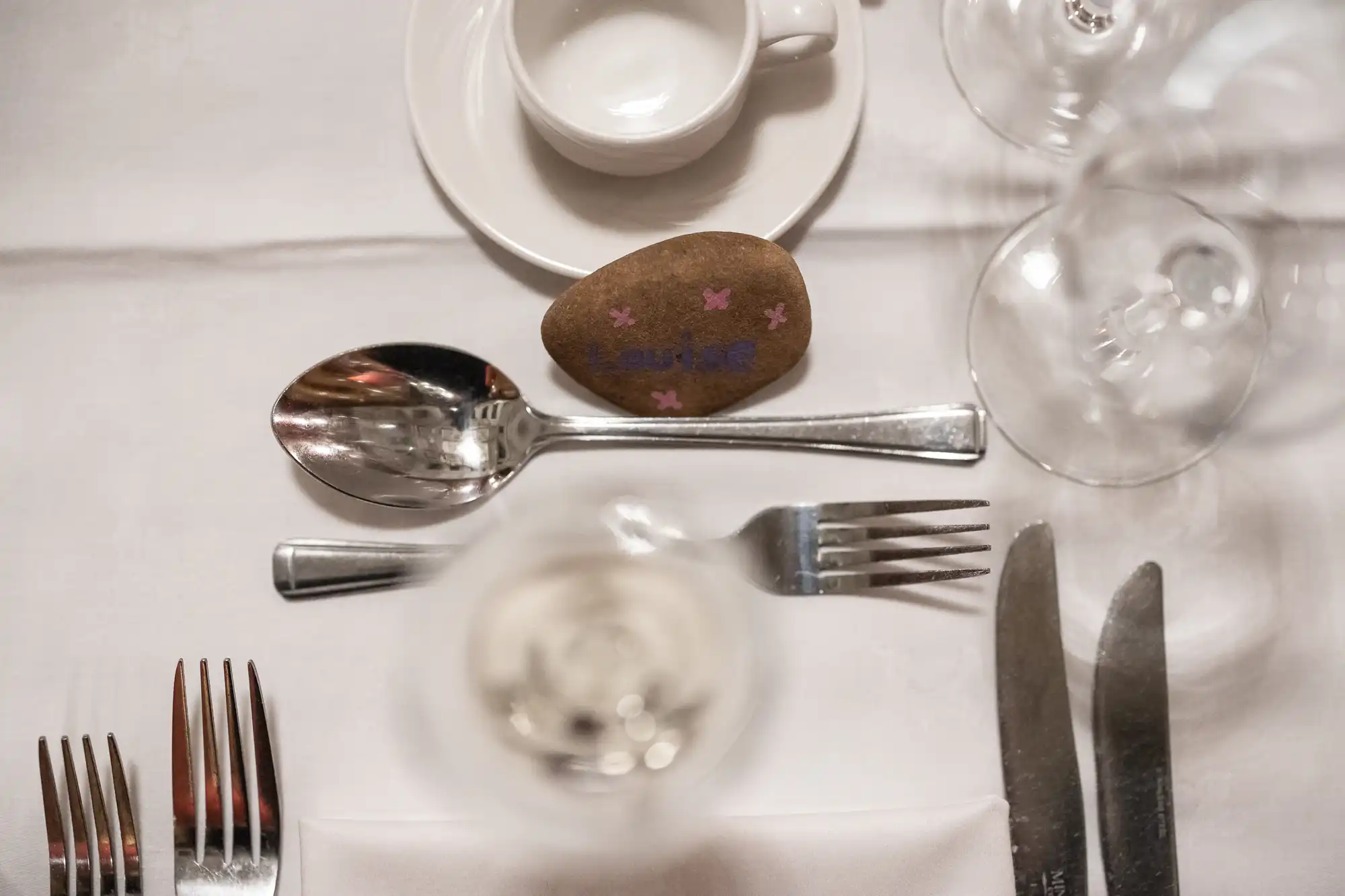Table setting with plates, glasses, silverware, a cup on a saucer, and a stone with purple markings and text on a white tablecloth.