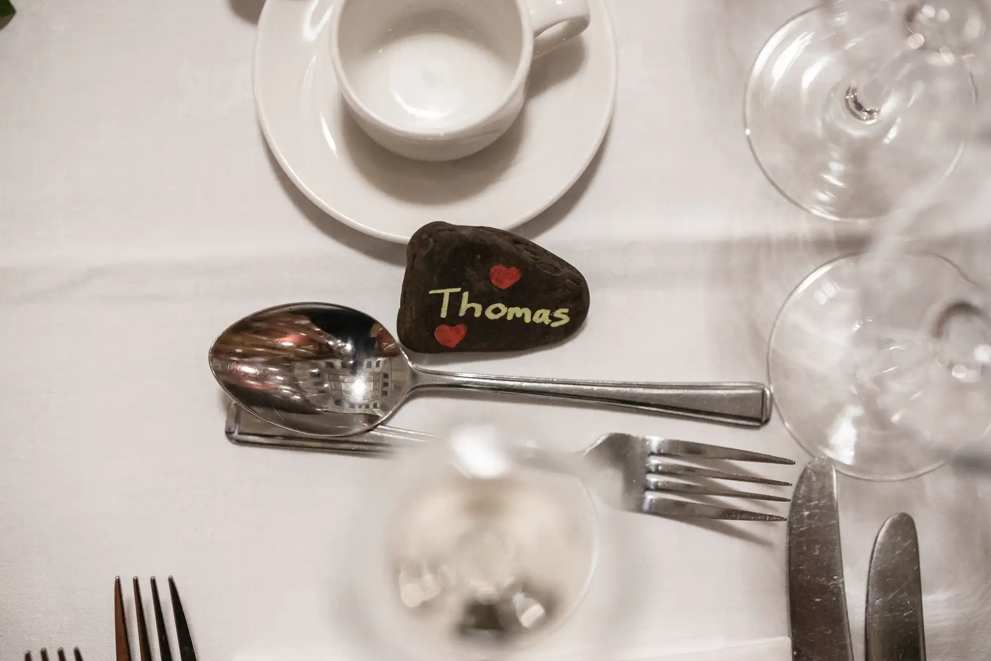 A place setting with a teacup, spoon, fork, and knife on a white tablecloth. A rock with "Thomas" and two red hearts painted on it is placed near the spoon.