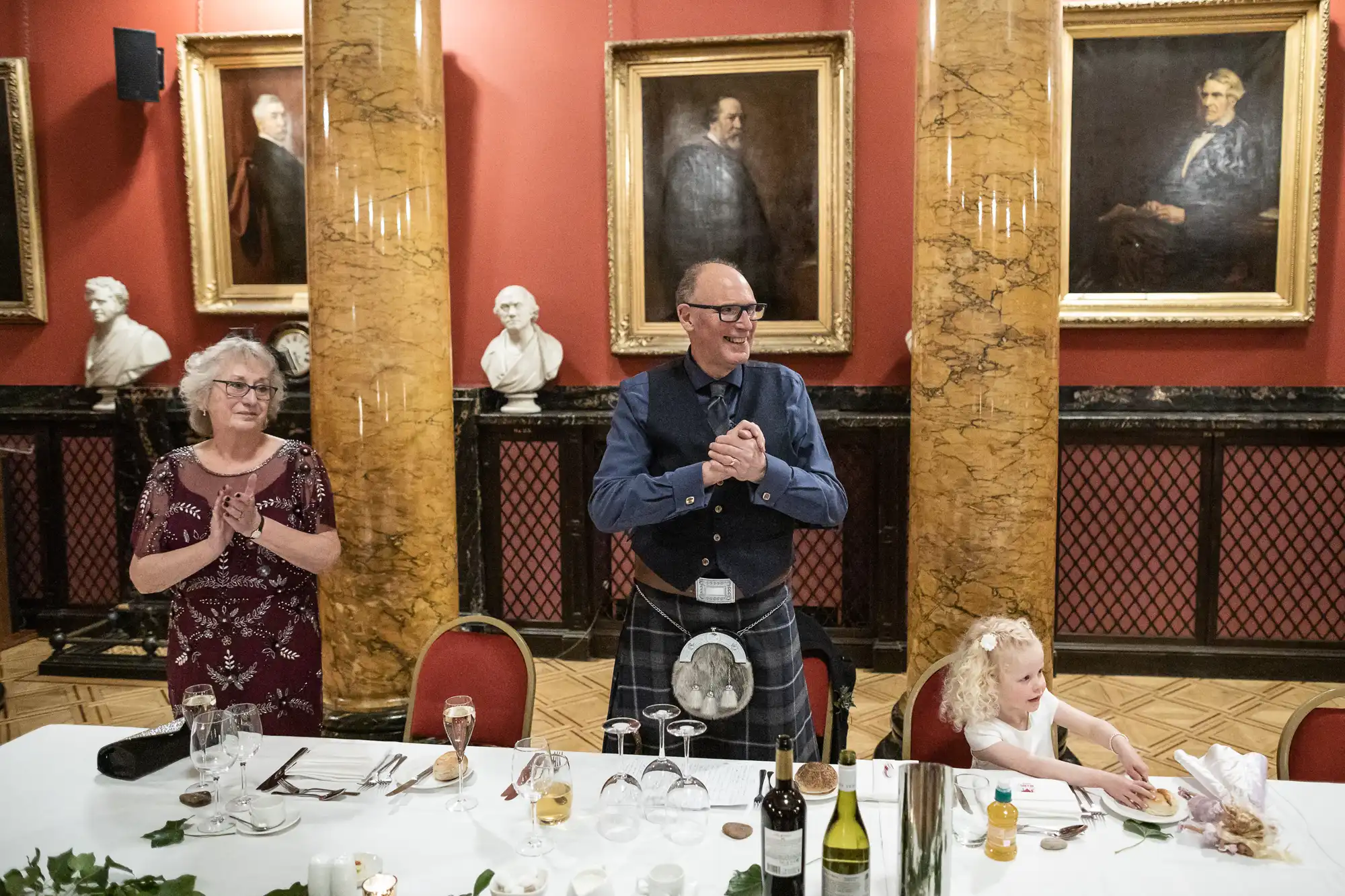 An elderly man in a kilt stands clapping between an elderly woman and a young girl seated at a table with food and drinks in a room decorated with portraits and marble columns.