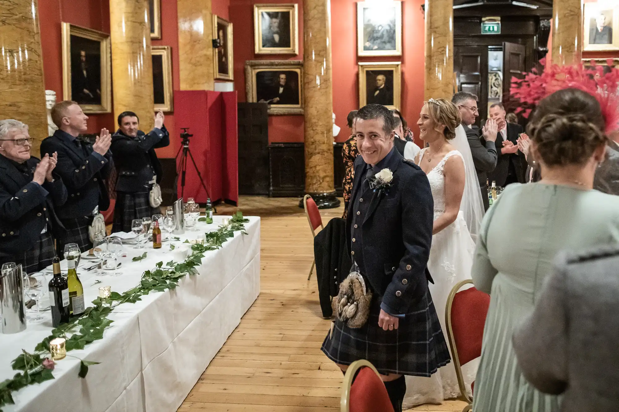 A groom in a kilt and a bride are greeted with applause as they walk into a reception in a hall with red walls and portraits. Guests are seated at decorated tables.