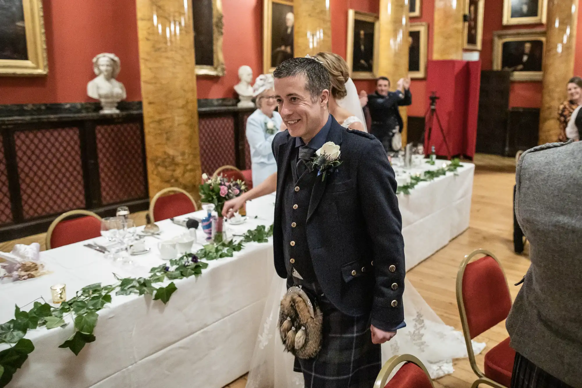 A man in traditional Scottish attire with a sporran walks among tables adorned with greenery and white linens in a formal setting, surrounded by guests.