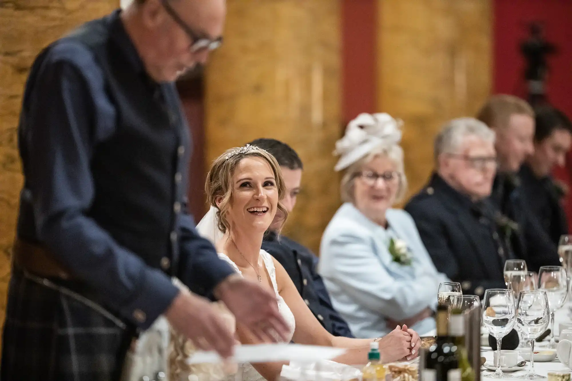 A bride sits smiling at a reception table, surrounded by guests. An older man stands and appears to be speaking, while other guests watch and listen.