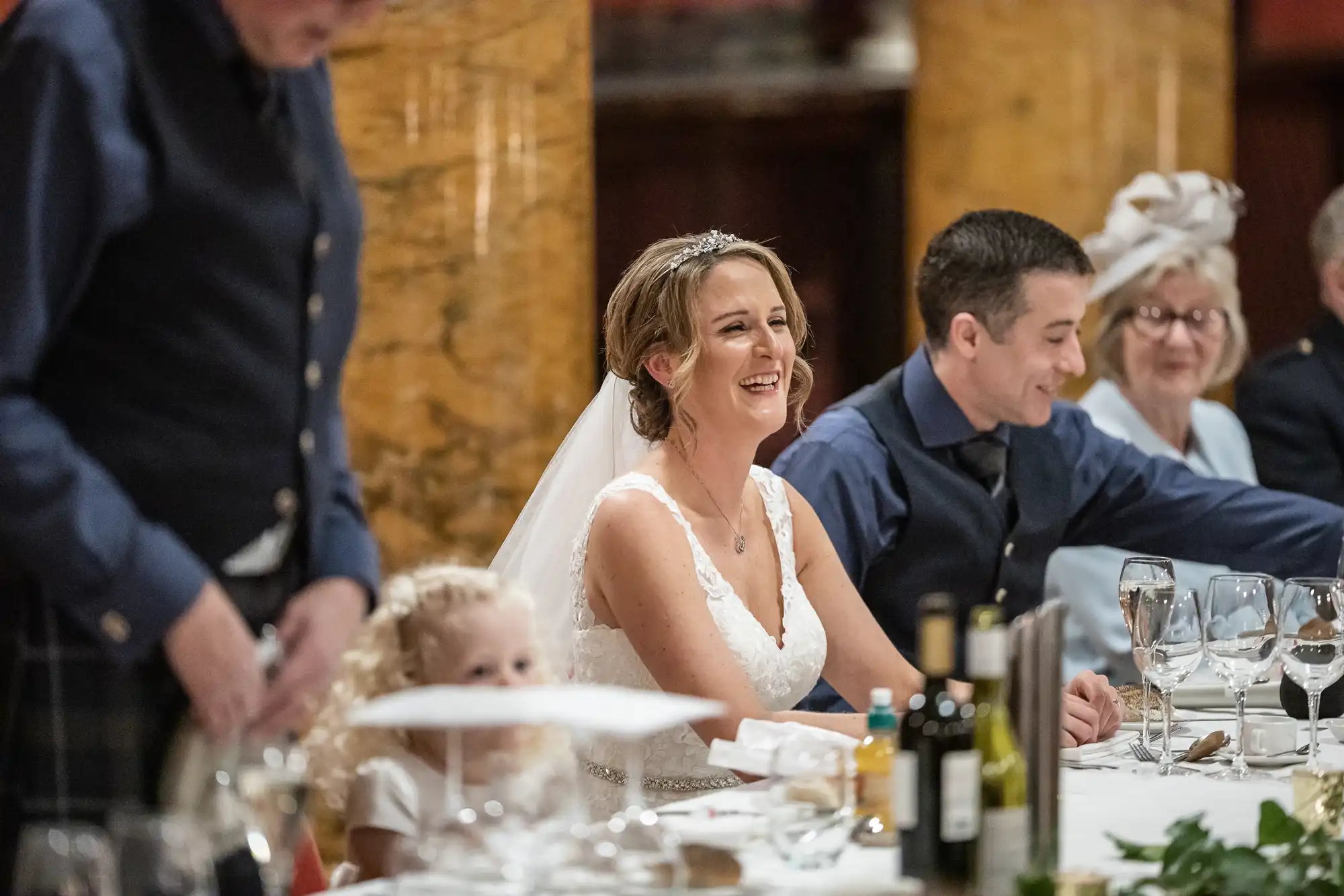 Bride in white dress smiles while seated at a dinner table with family and friends, including a child in the foreground and guests wearing formal attire.
