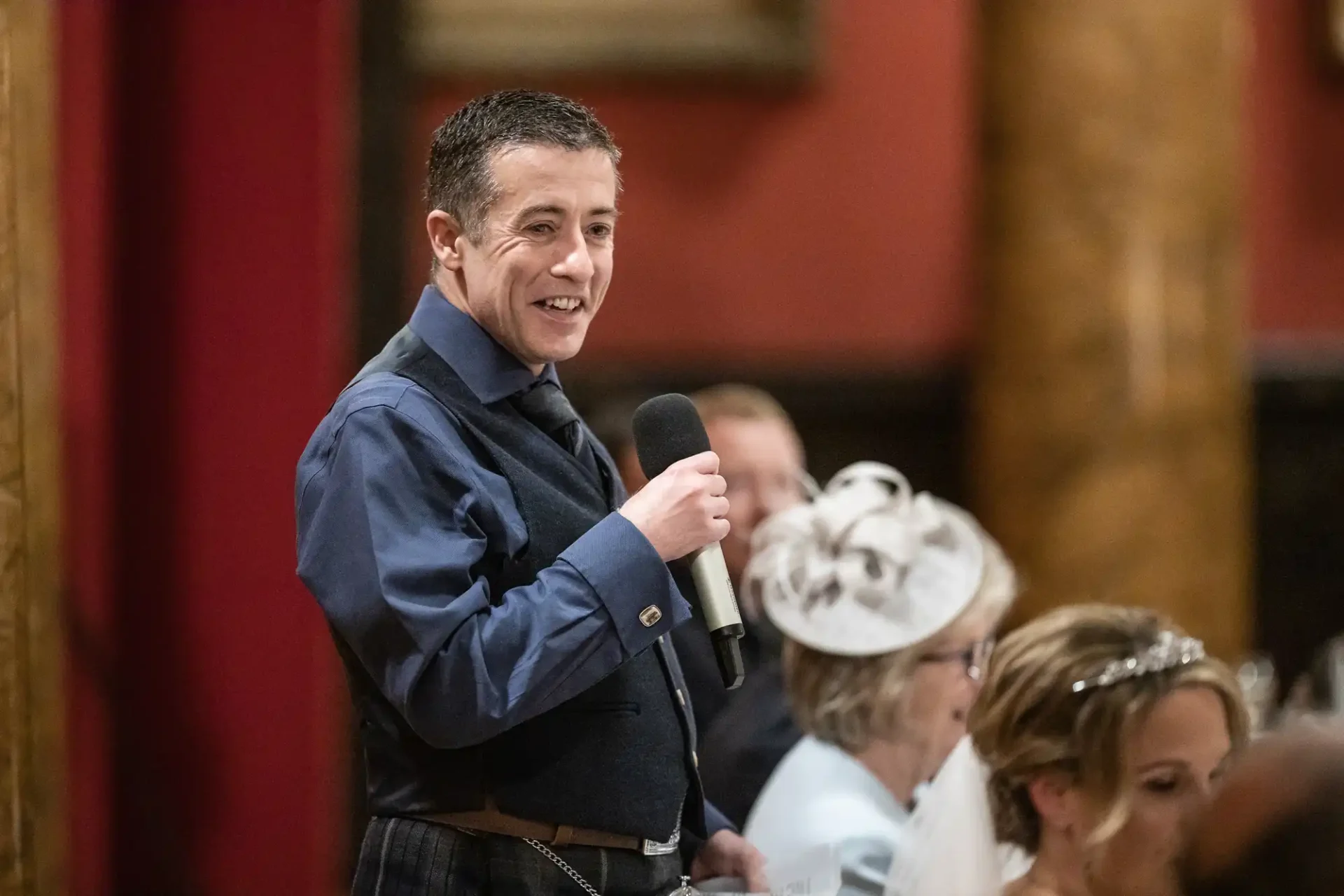A man in a dark vest and shirt holds a microphone while speaking at an indoor event. Others seated in the background are dressed formally.