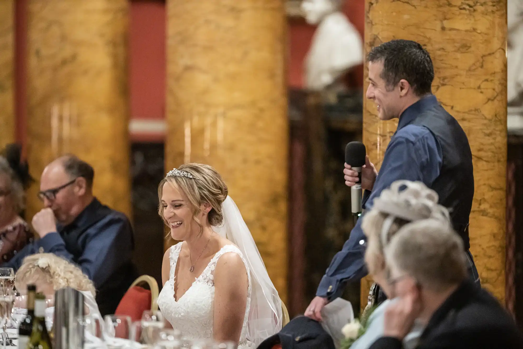 A man speaks into a microphone while a woman in a white dress and veil smiles, surrounded by seated guests in a room with large, yellow marble columns.