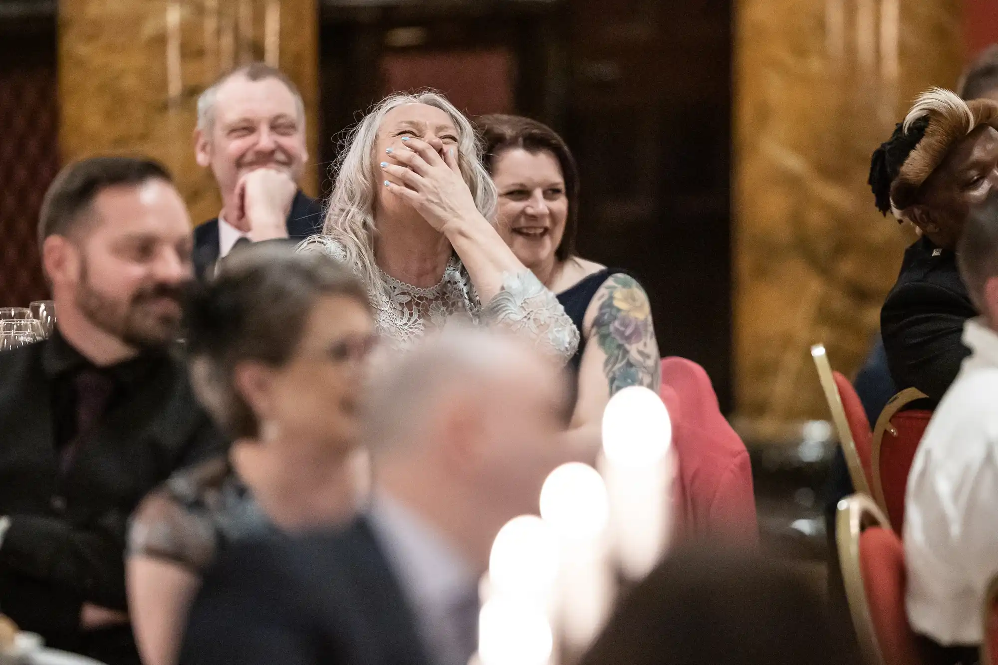 A group of people are seated at a formal event. A woman in focus is laughing with her hand covering her mouth, while others around her are smiling.