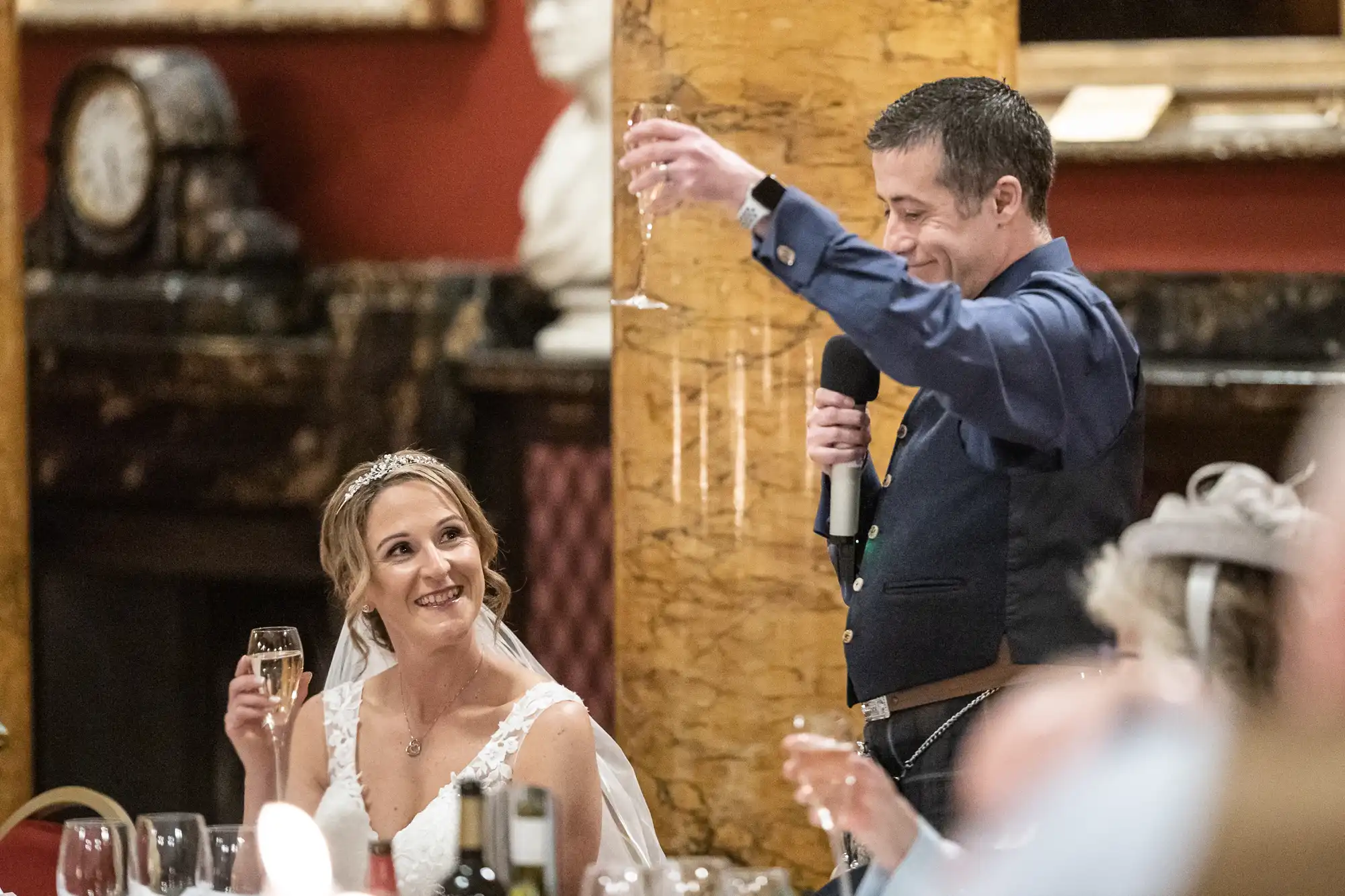 A groom gives a speech holding a microphone and champagne flute, while the bride in a white dress and veil looks at him smiling, sitting at a table with bottles and glasses.