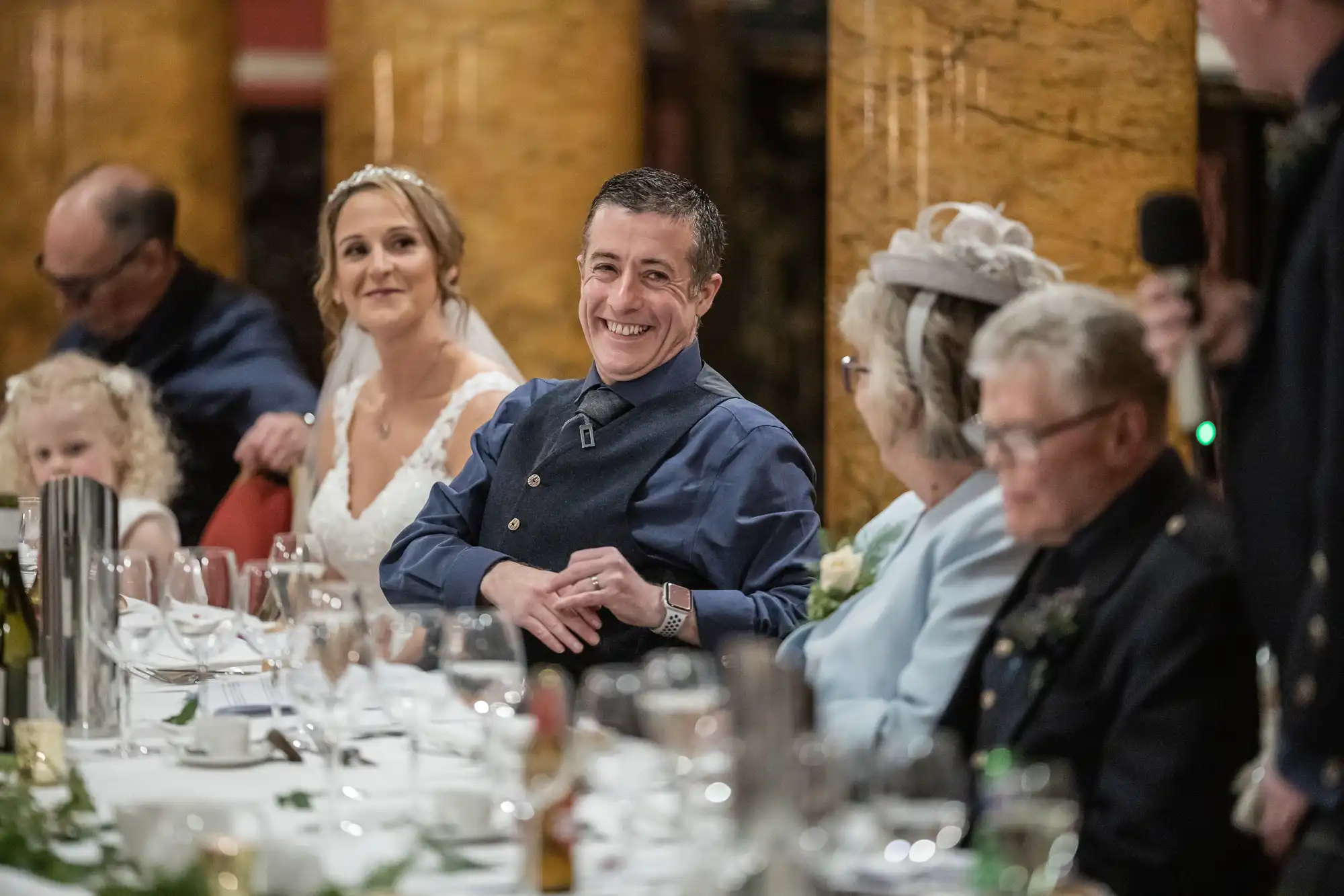 A wedding reception scene with a bride, groom, and guests seated at a long table. The groom smiles, and the bride looks on with other family members and friends.