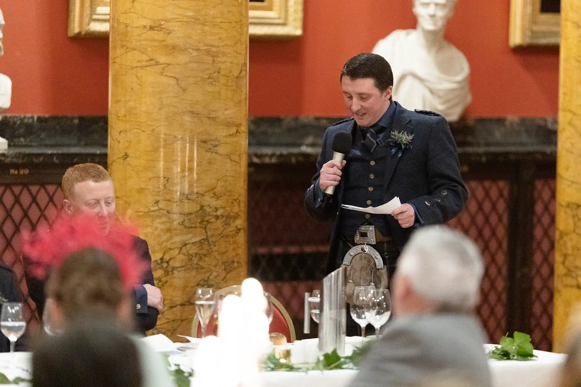 A man in traditional Scottish attire speaks into a microphone while holding a piece of paper at a formal event with seated guests.
