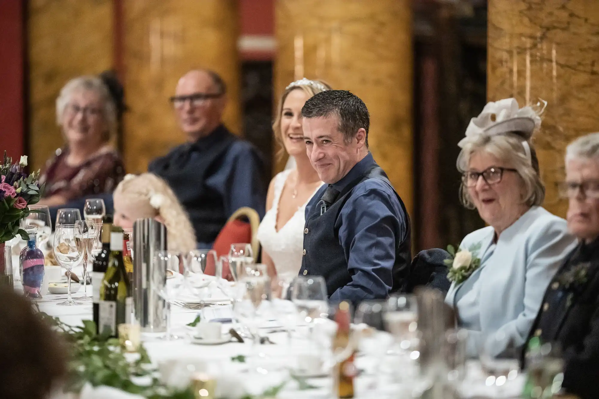A group of people dressed formally are seated at a long table with white tablecloth, glassware, and plates. Prominently, a bride and man in a blue vest smile while others converse and look on.