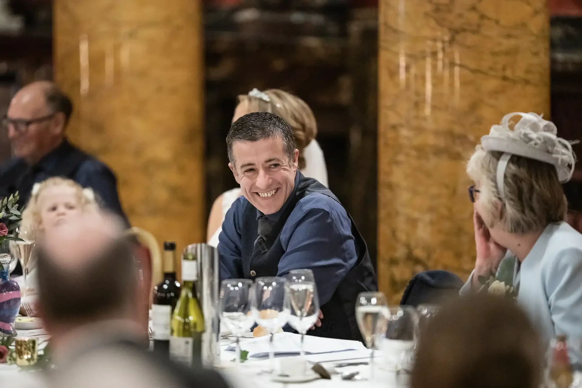 Several people sit around a table adorned with wine glasses and bottles, with one person smiling warmly at another during what appears to be a social event in an elegantly decorated venue.