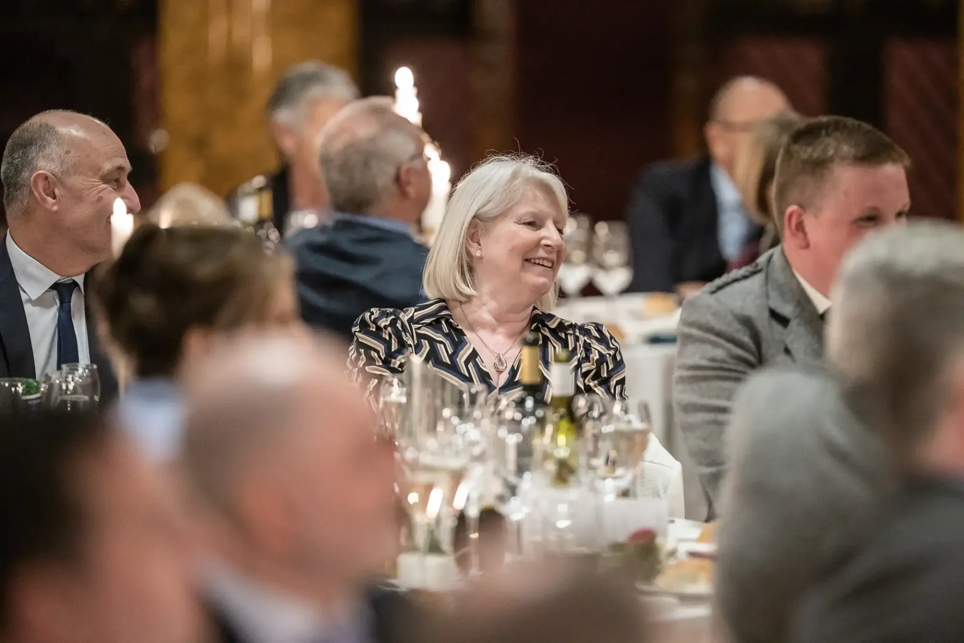 People seated at a formal event. A woman at the center is smiling, surrounded by others dressed in business attire. The atmosphere appears to be professional and convivial.