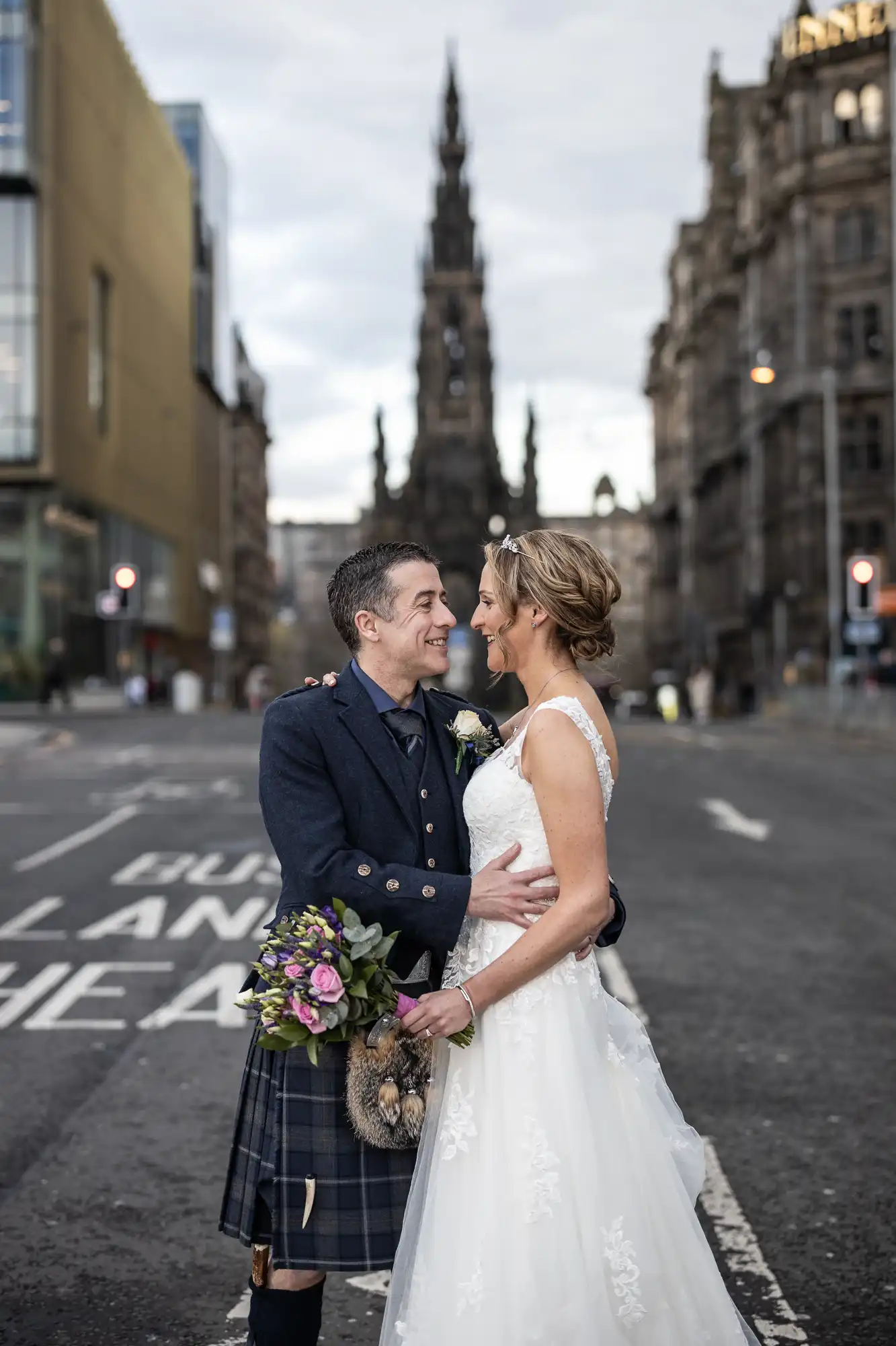 A couple wearing wedding attire, with the groom in a kilt and the bride in a white gown, stand embracing on a city street with historic buildings and a tall monument in the background.