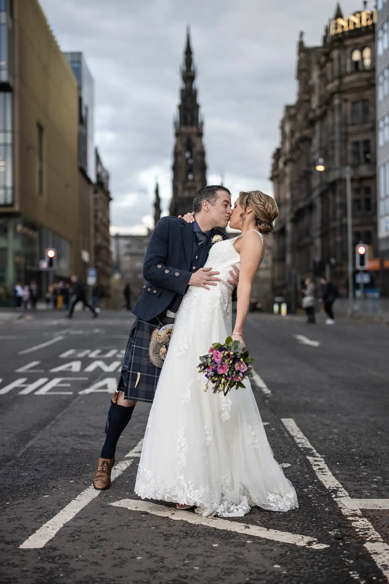 A bride and groom kiss on an urban street, with the bride holding a bouquet and the groom wearing a kilt. A tall monument and historic buildings are visible in the background.