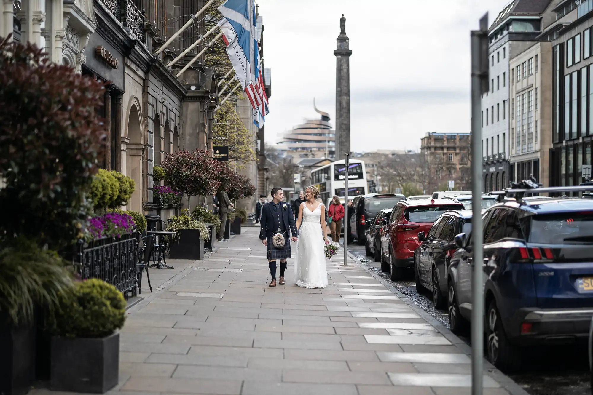 A couple in wedding attire walks down a city sidewalk, with buildings, parked cars, and flags in the background.