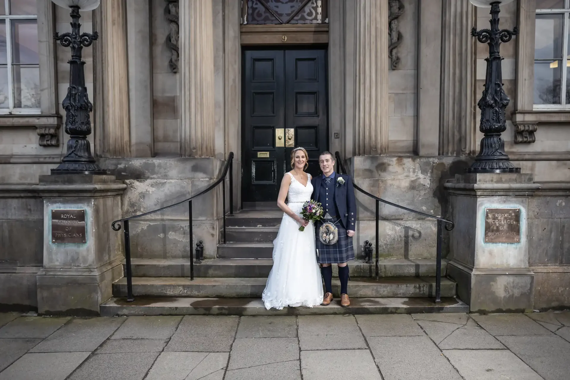 A bride in a white dress and a groom in a kilt stand side by side on the steps of an old building with large double doors and stone pillars.