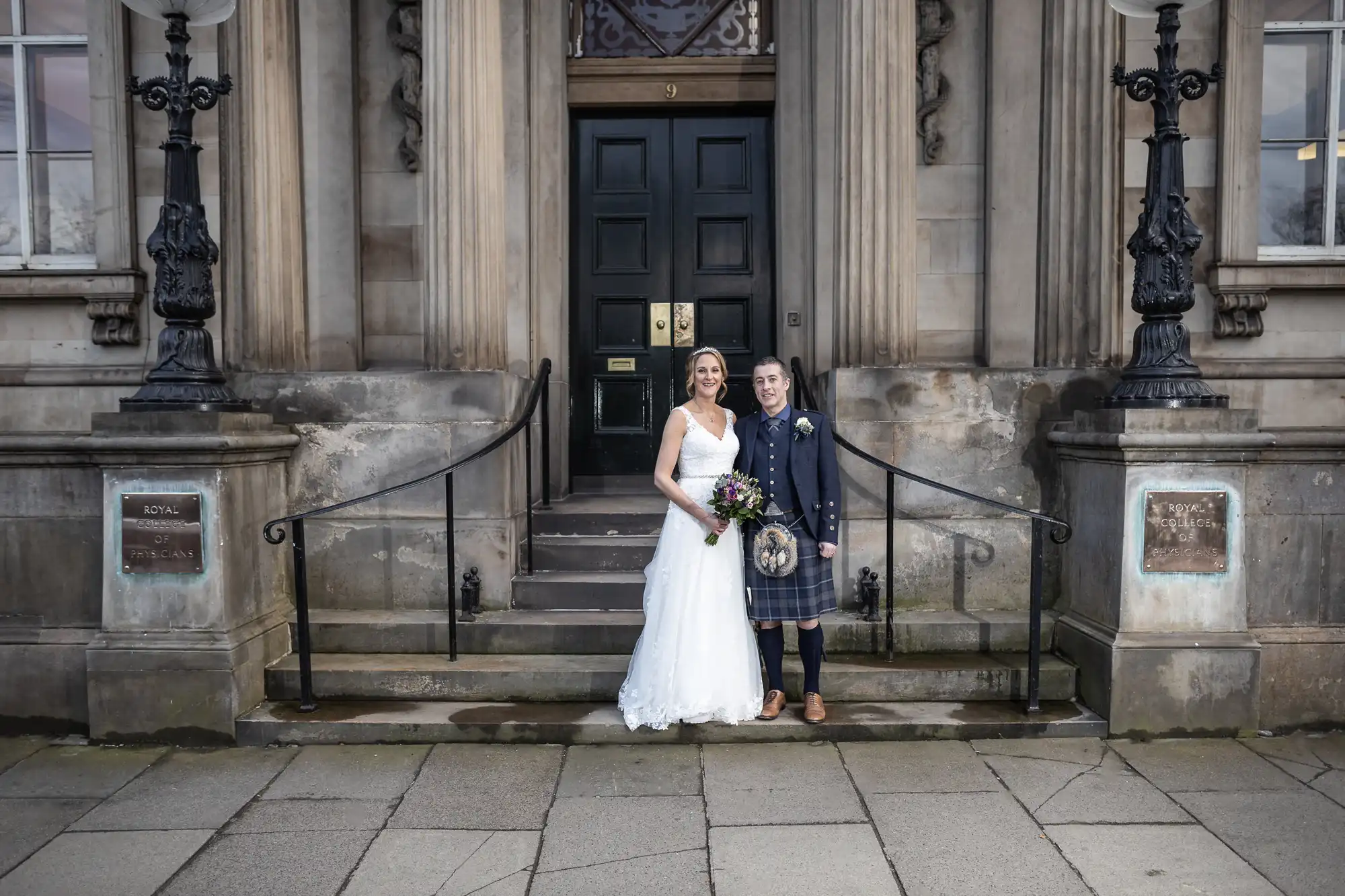 A bride in a white dress and a groom in a kilt stand side by side on the steps of an old building with large double doors and stone pillars.