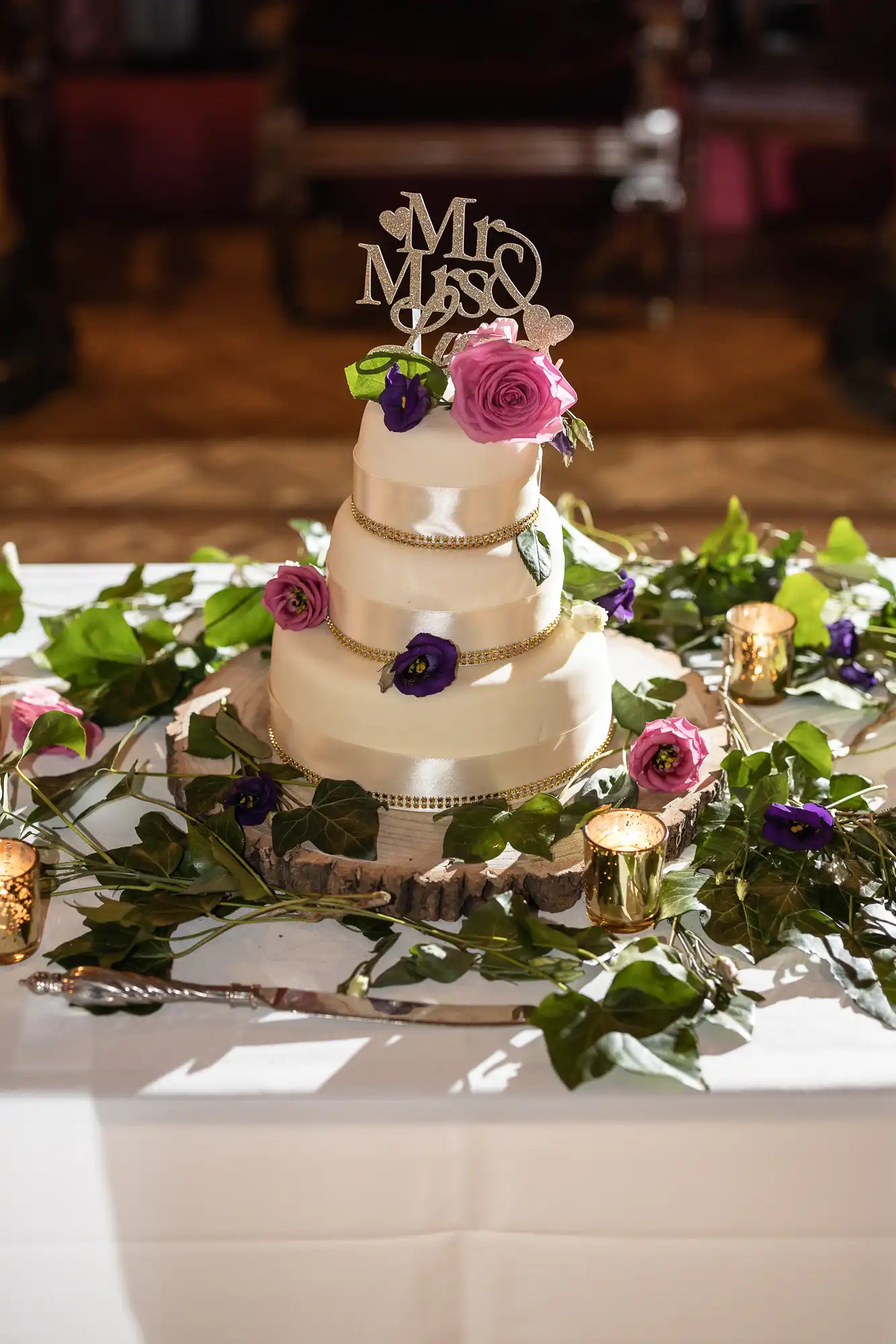 A three-tiered white wedding cake is decorated with purple flowers, greenery, and a "Mr & Mrs" topper. The cake is placed on a white table adorned with foliage and candles.