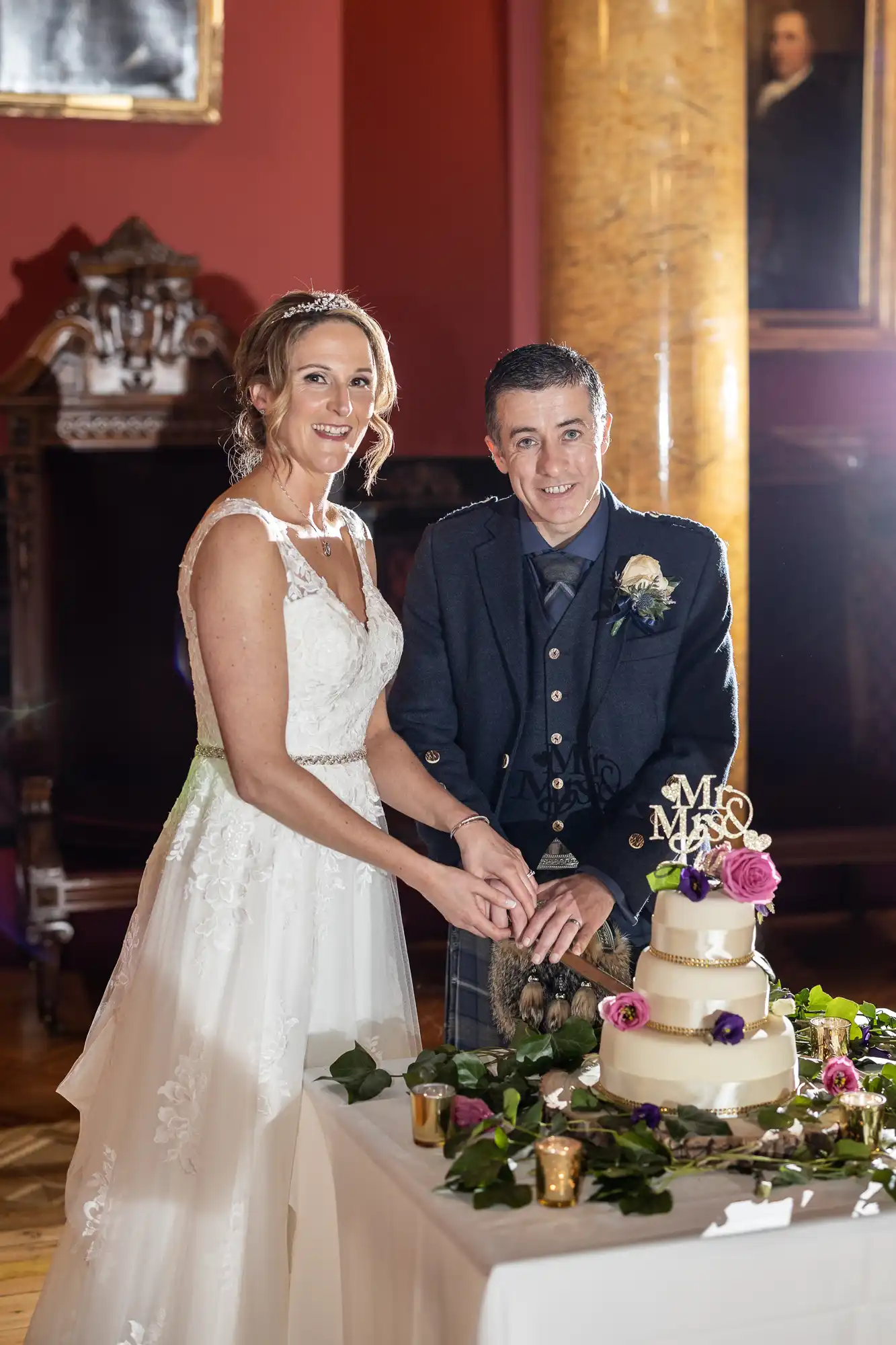 A bride and groom in wedding attire smile as they cut a tiered wedding cake decorated with flowers and a "Mr & Mrs" topper. They stand in an ornate room with a column and framed painting in the background.