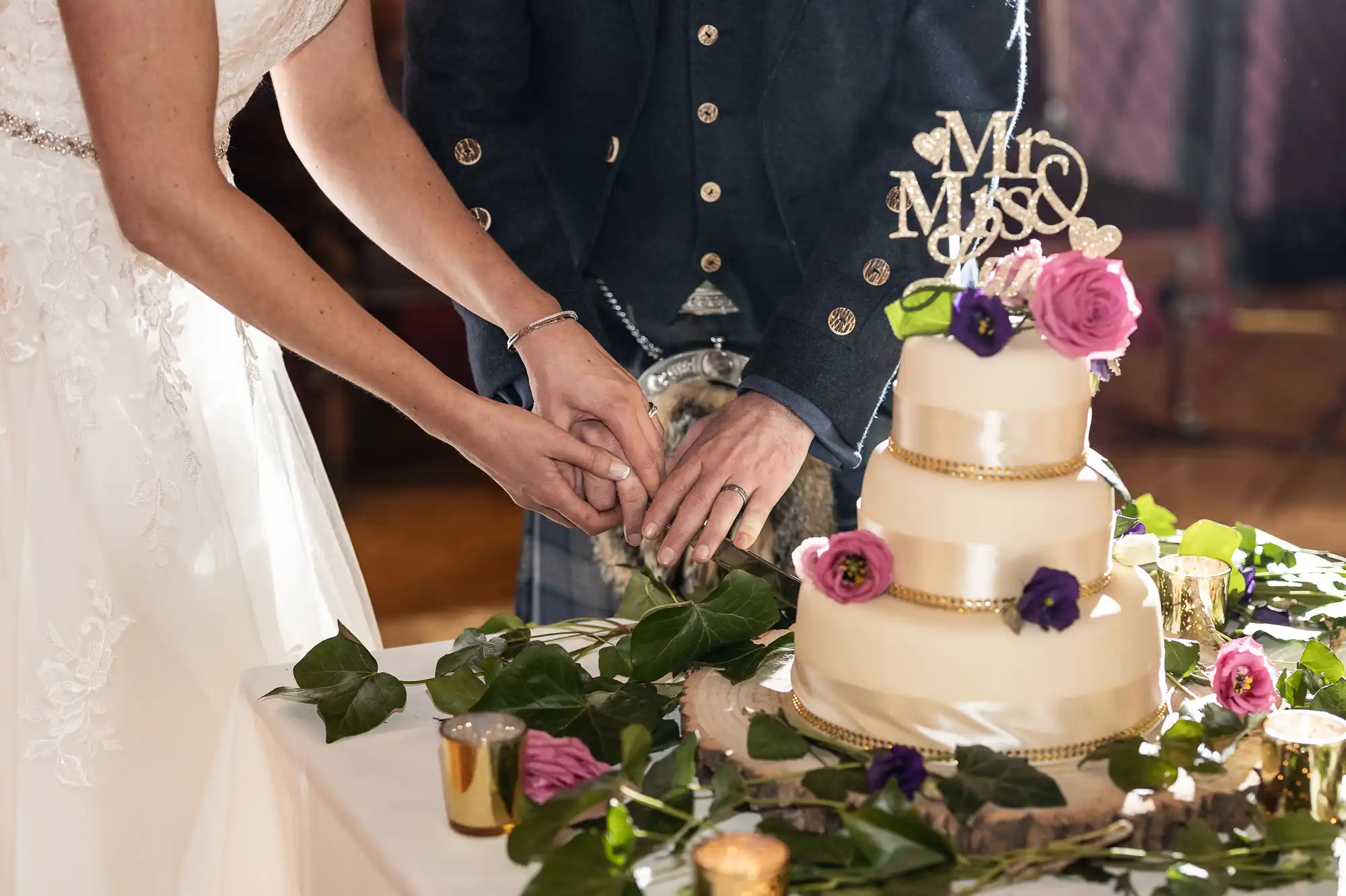 A bride and groom cut into a three-tier wedding cake adorned with flowers and a "Mr & Mrs" topper, standing on a table decorated with foliage and candles.