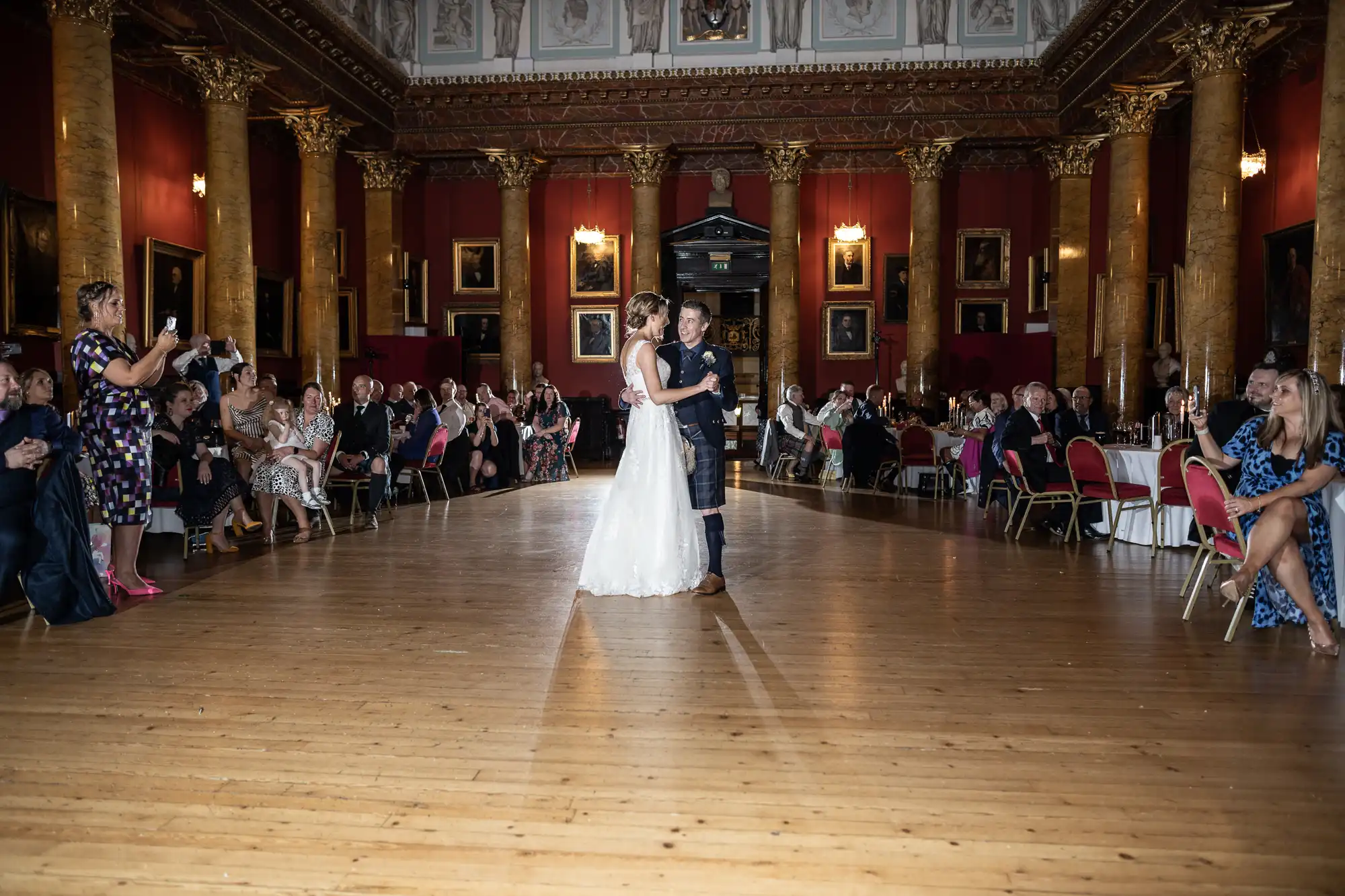 A bride and groom are dancing in a grand hall with guests seated on either side, watching and taking photos. The room features ornate columns, red walls, and framed paintings.