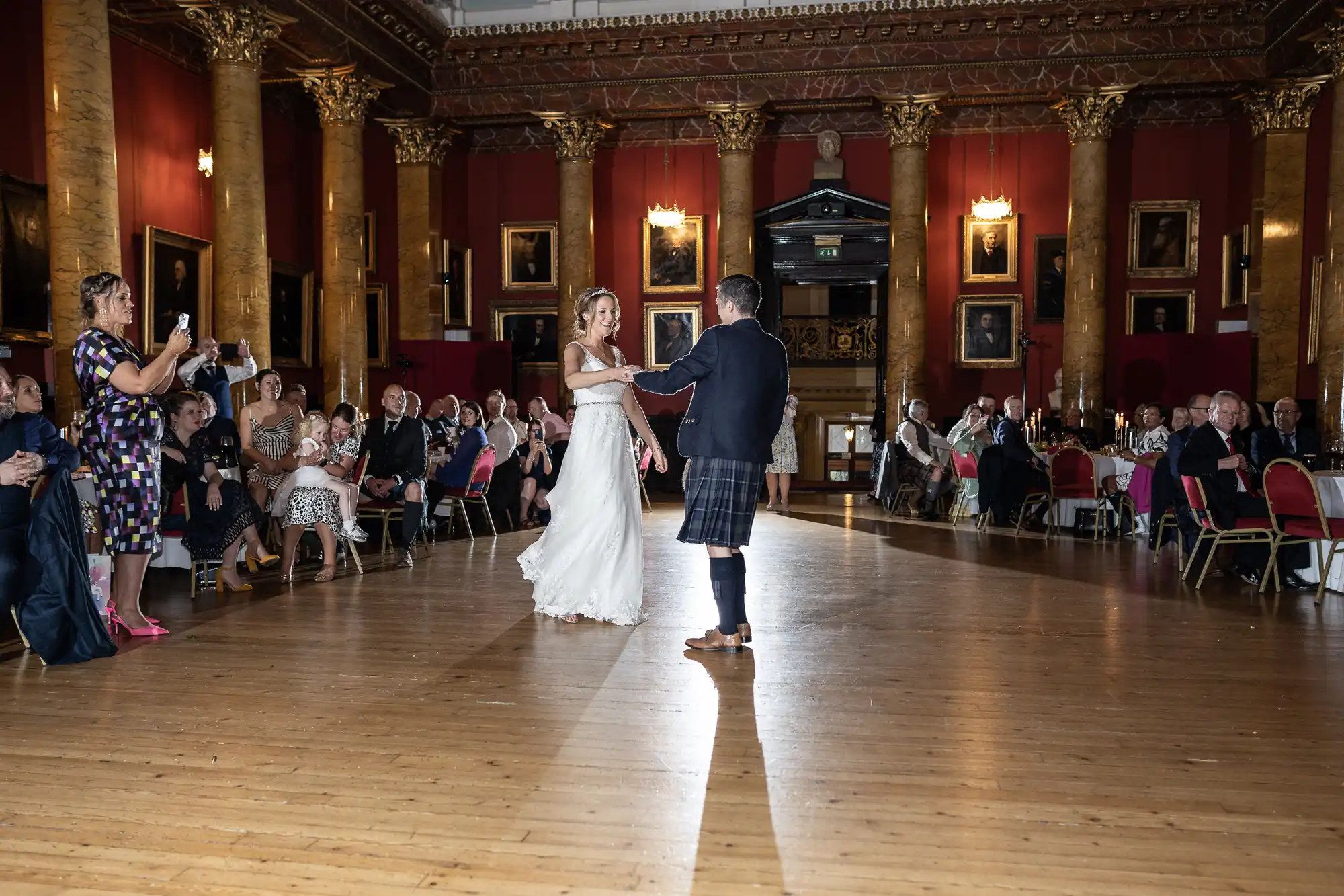 A bride and groom dance in an ornate hall with wooden floors, watched by seated guests. The room features tall columns and framed portraits on the walls.