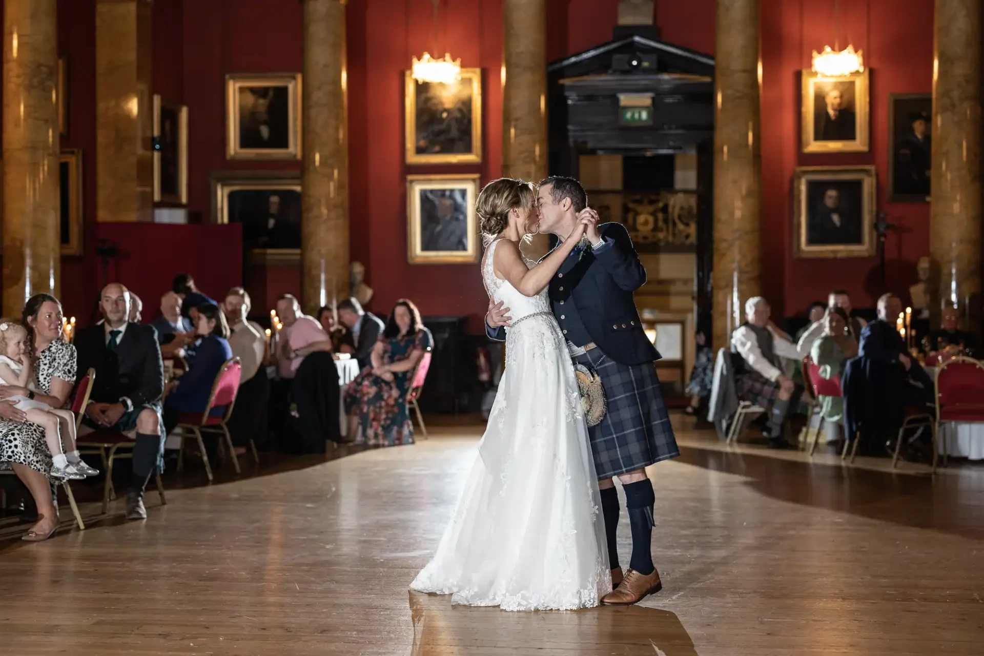 A couple in formal attire shares a first dance in an elegant hall with guests seated at tables in the background.