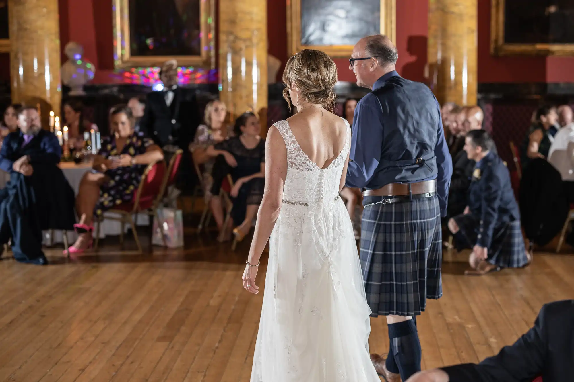 A couple wearing formal attire, including a white dress and a kilt, walk across a wooden floor in a dimly lit room with seated guests in the background.