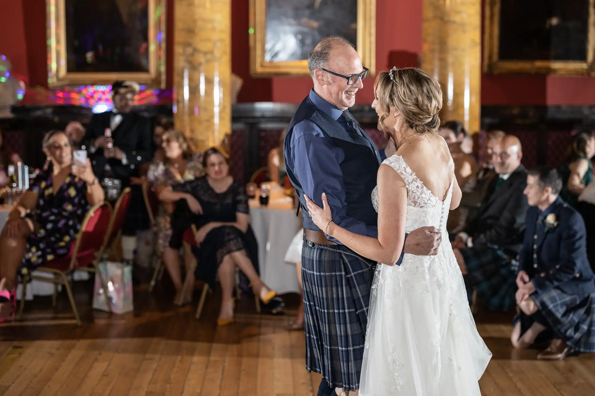 A couple dances while guests sit and watch in a warmly lit room decorated with framed paintings and dark wood panels. The woman wears a white dress, and the man wears a kilt and dark shirt.