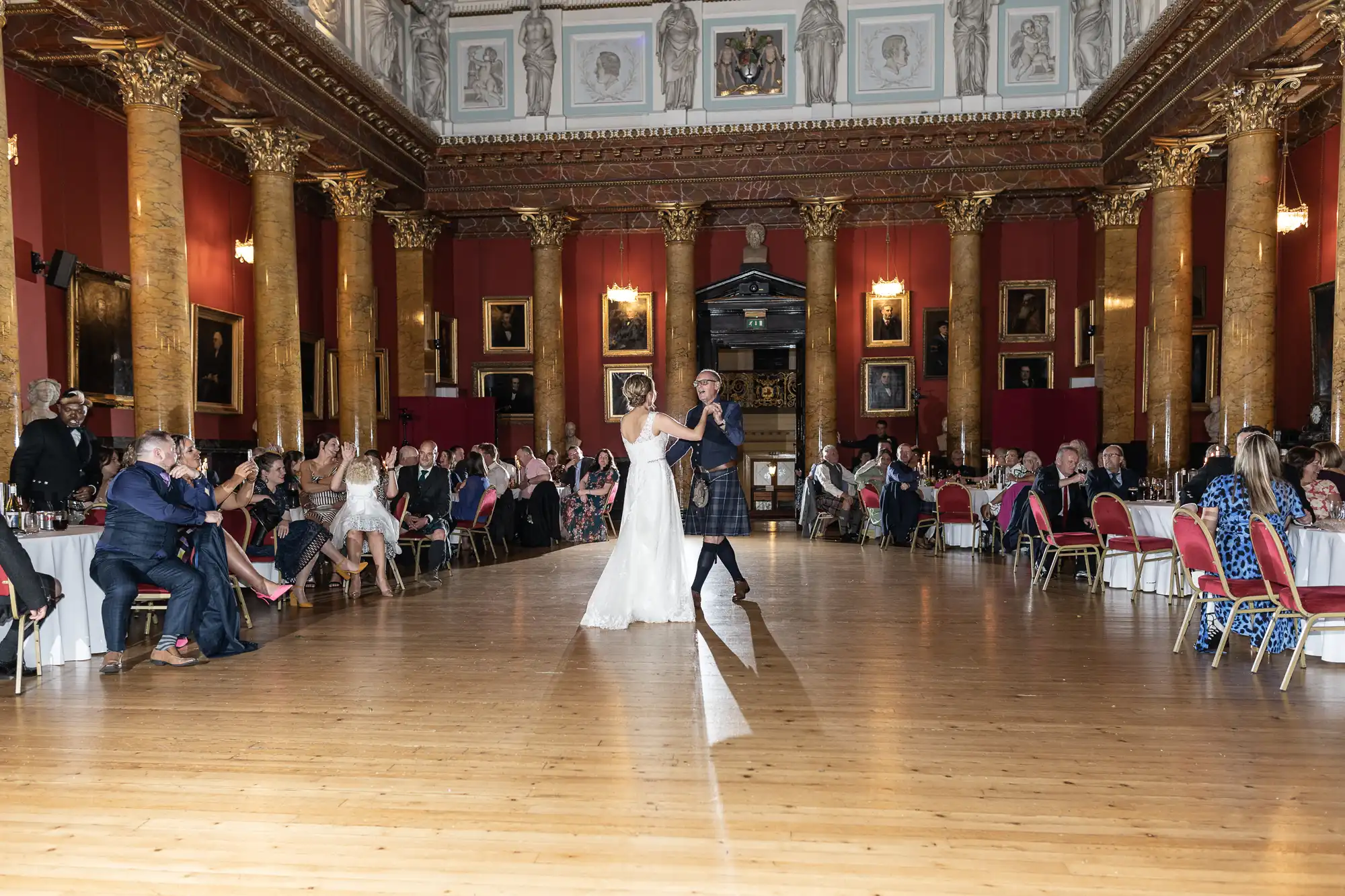 A couple in formal attire, a wedding gown, and a kilt, dances in the center of a large, ornate hall with guests seated around the edges, watching.