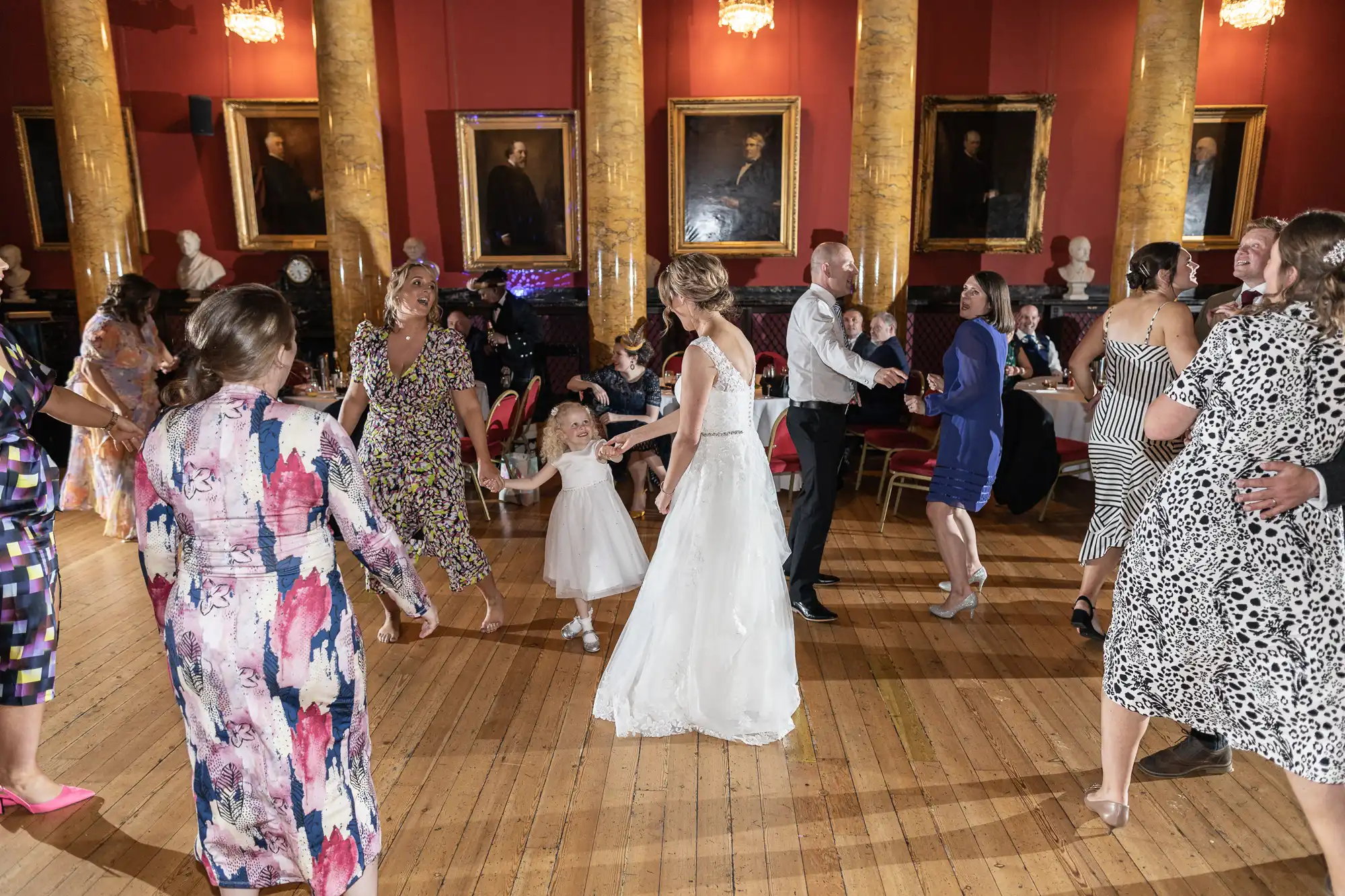 Guests, including a bride in a white dress, are dancing in a large hall with wooden floors and portraits on the walls. There is a little girl in a white dress holding hands with the bride.