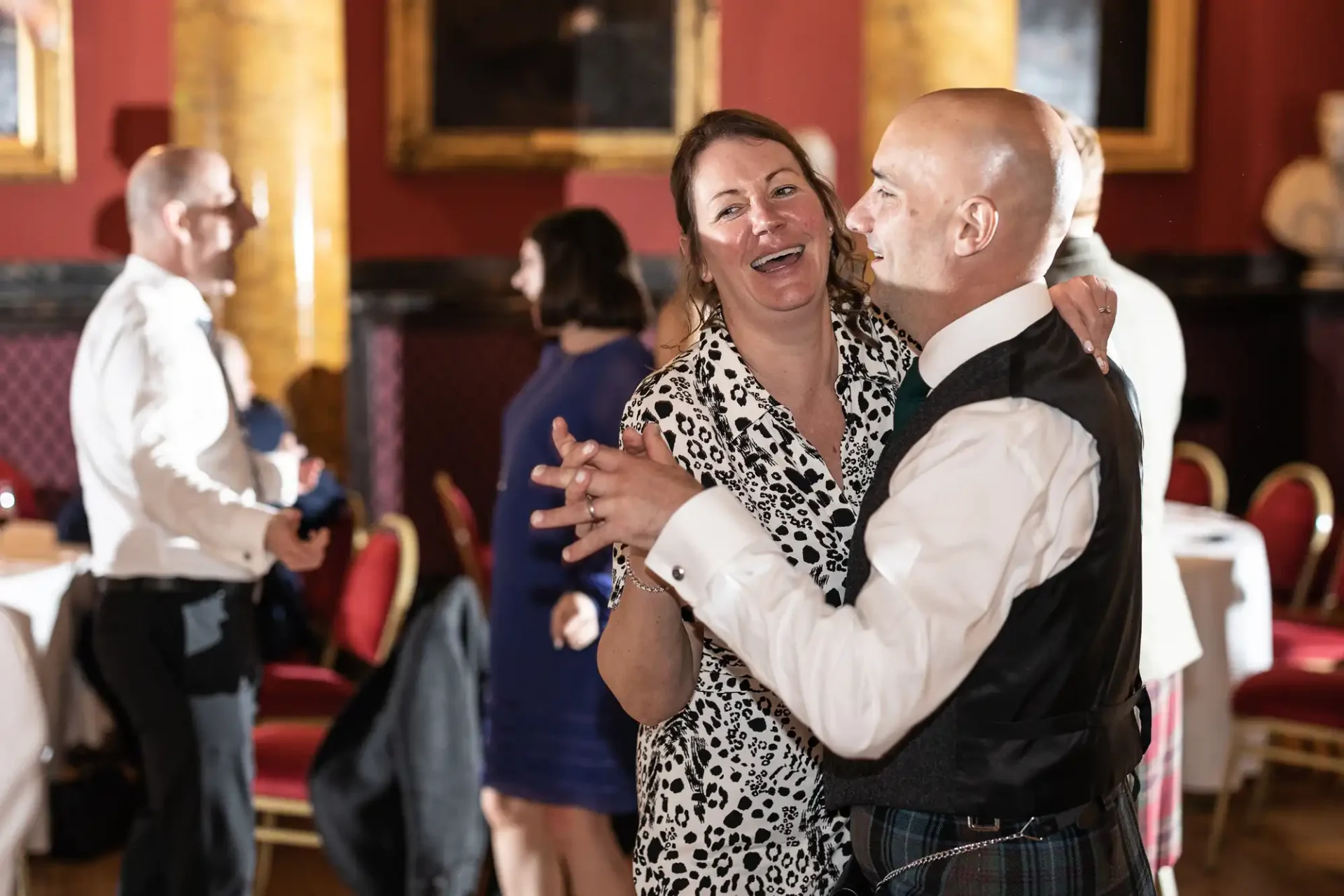 A man and a woman dance together at an event, smiling. Other attendees in formal attire are visible in the background.