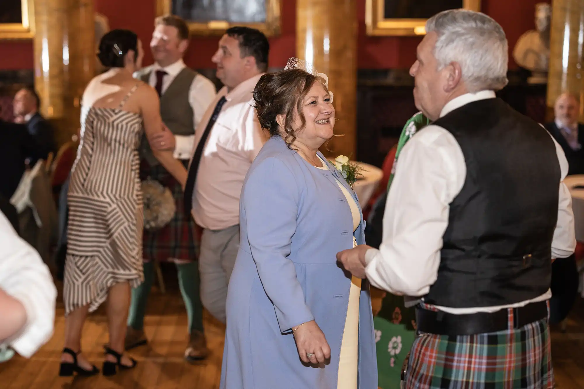 A group of people are dancing in an elegant room with red walls and columns. One woman in a blue coat and a man in a black vest and kilt are in the foreground.
