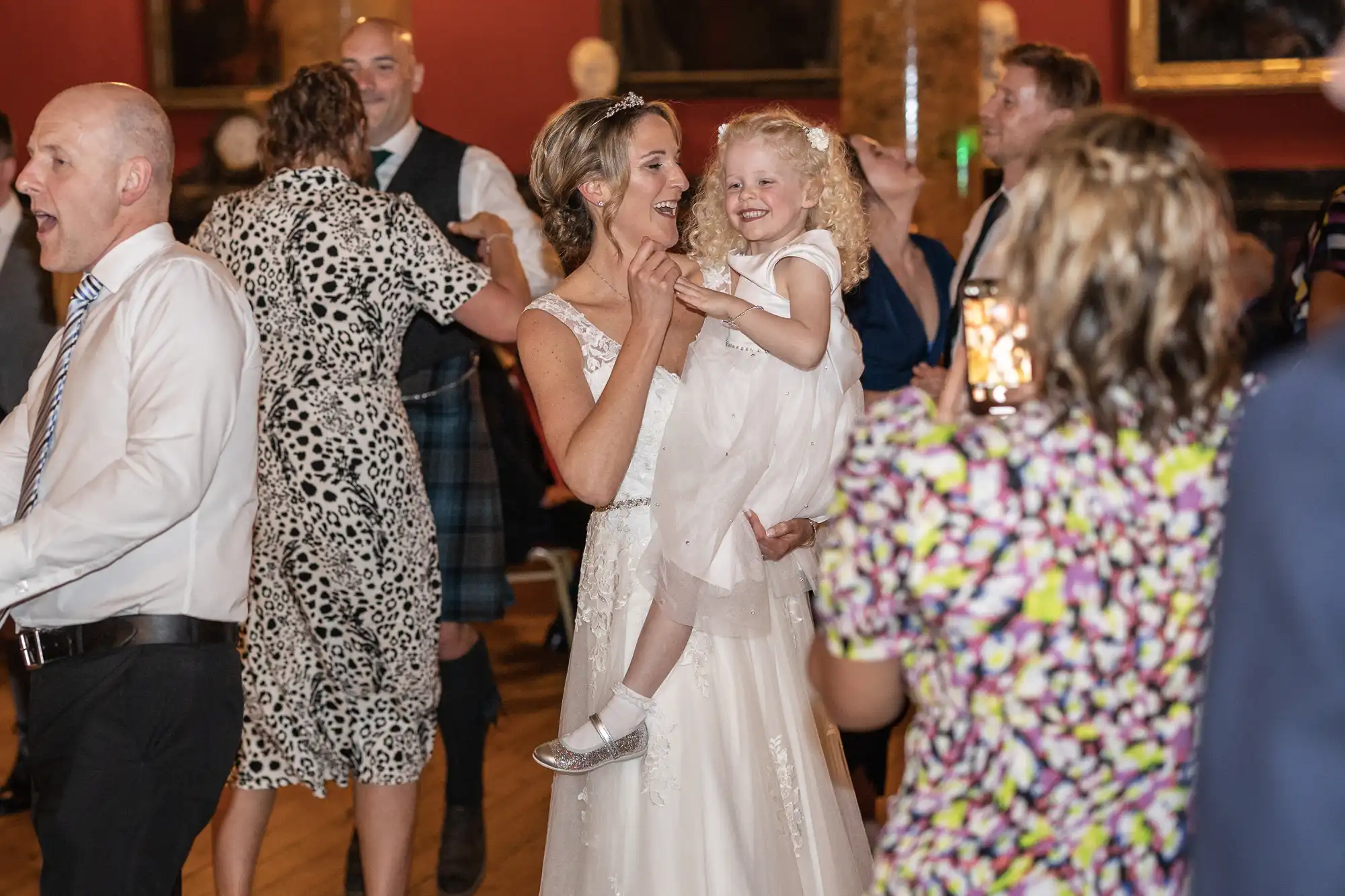 A bride holds a young girl in her arms while dancing at a wedding reception. Several other guests are also dancing around them.
