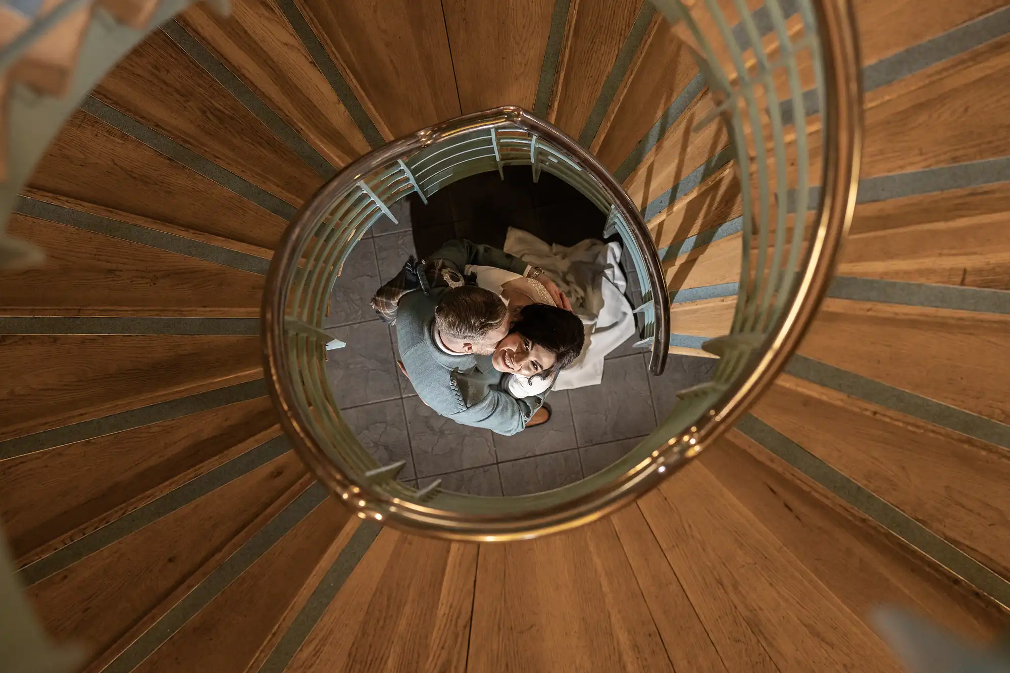 A couple embraces while standing at the bottom of a spiral staircase, viewed from above. The wooden steps and metal railing create a circular frame around them.