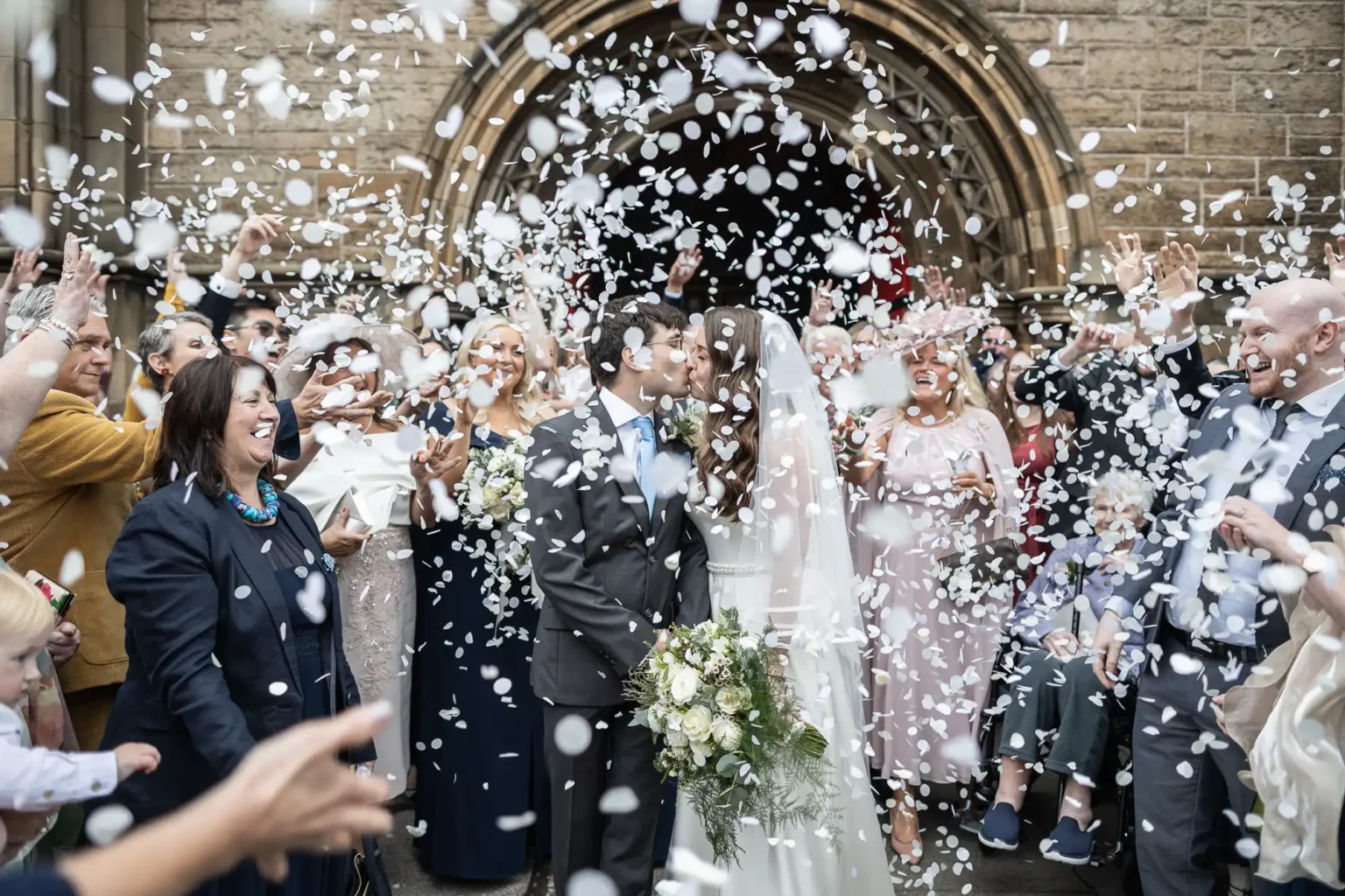 A newlywed couple kisses outside a church as guests stand around them, tossing white confetti in celebration.