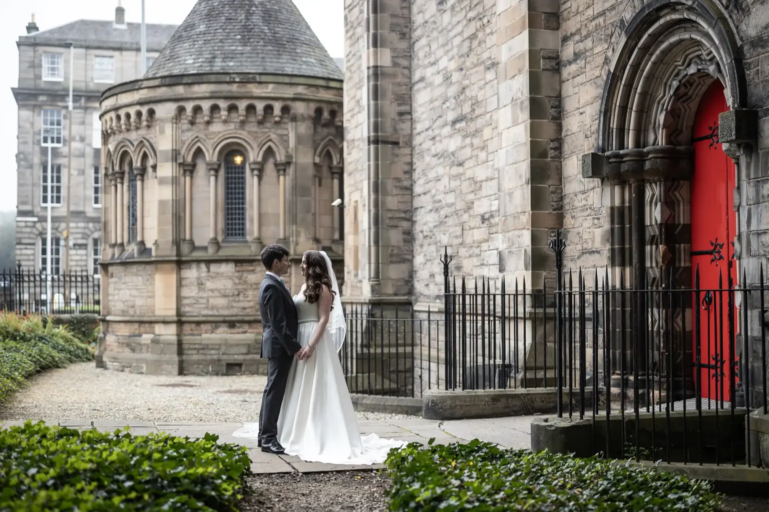 A bride and groom stand facing each other outside a historic stone church with arched windows and a red door at Mansfield Traquair, Edinburgh.