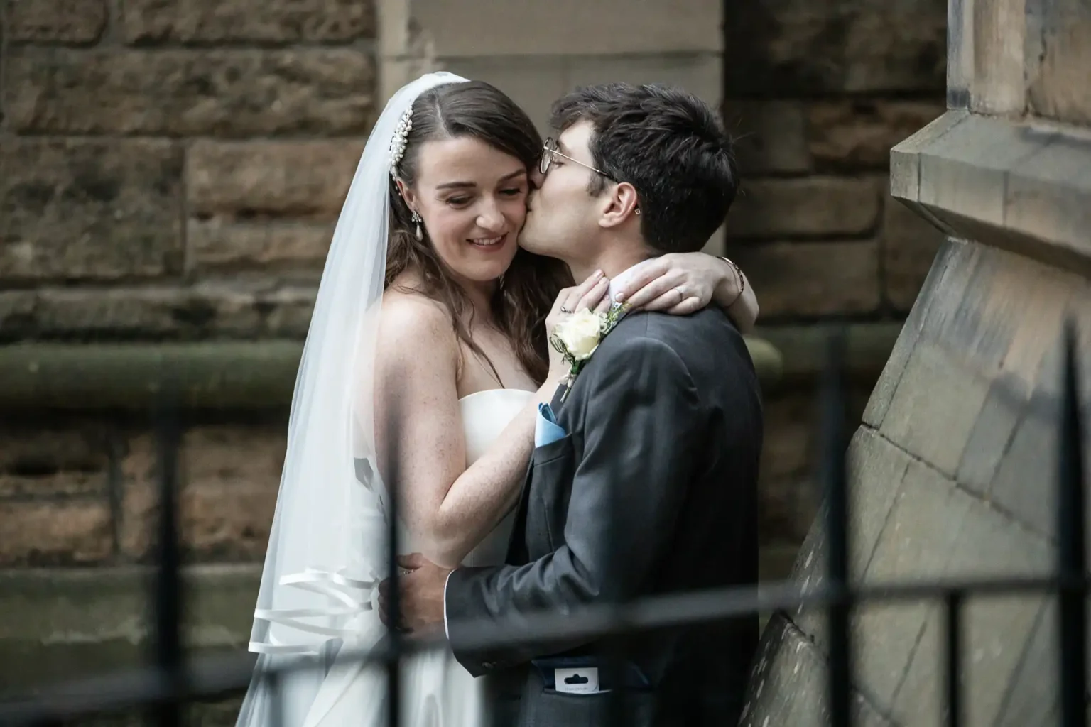 A bride in a white dress and veil smiles as a groom in a suit kisses her on the cheek outside a stone building.