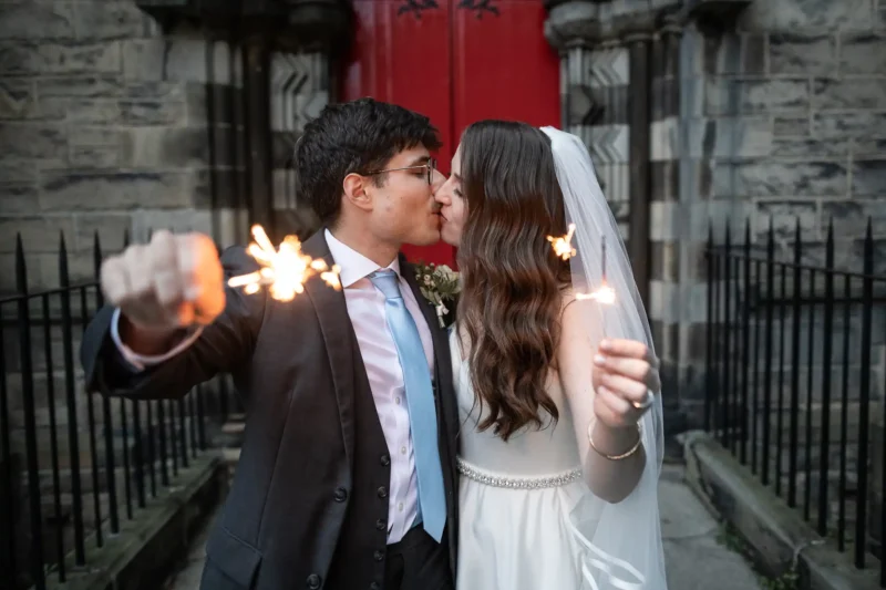 A bride and groom kiss while holding sparklers outside a building with stone walls and a red door.