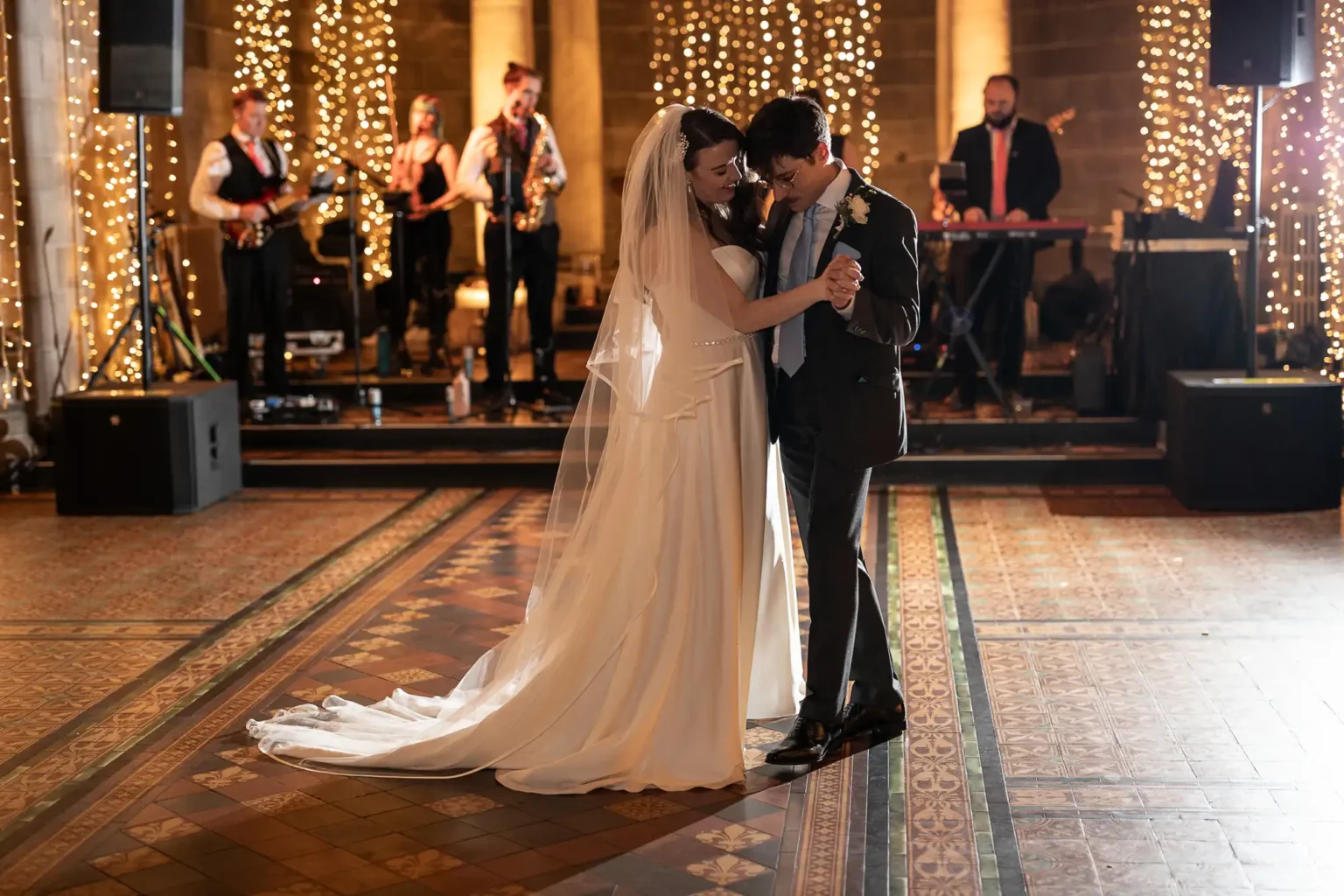 A bride and groom share their first dance in an elegant venue with string lights and a live band performing in the background.