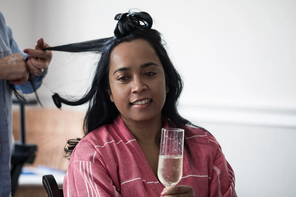 bride enjoys a glass of champagne while having her hair done pre-ceremony