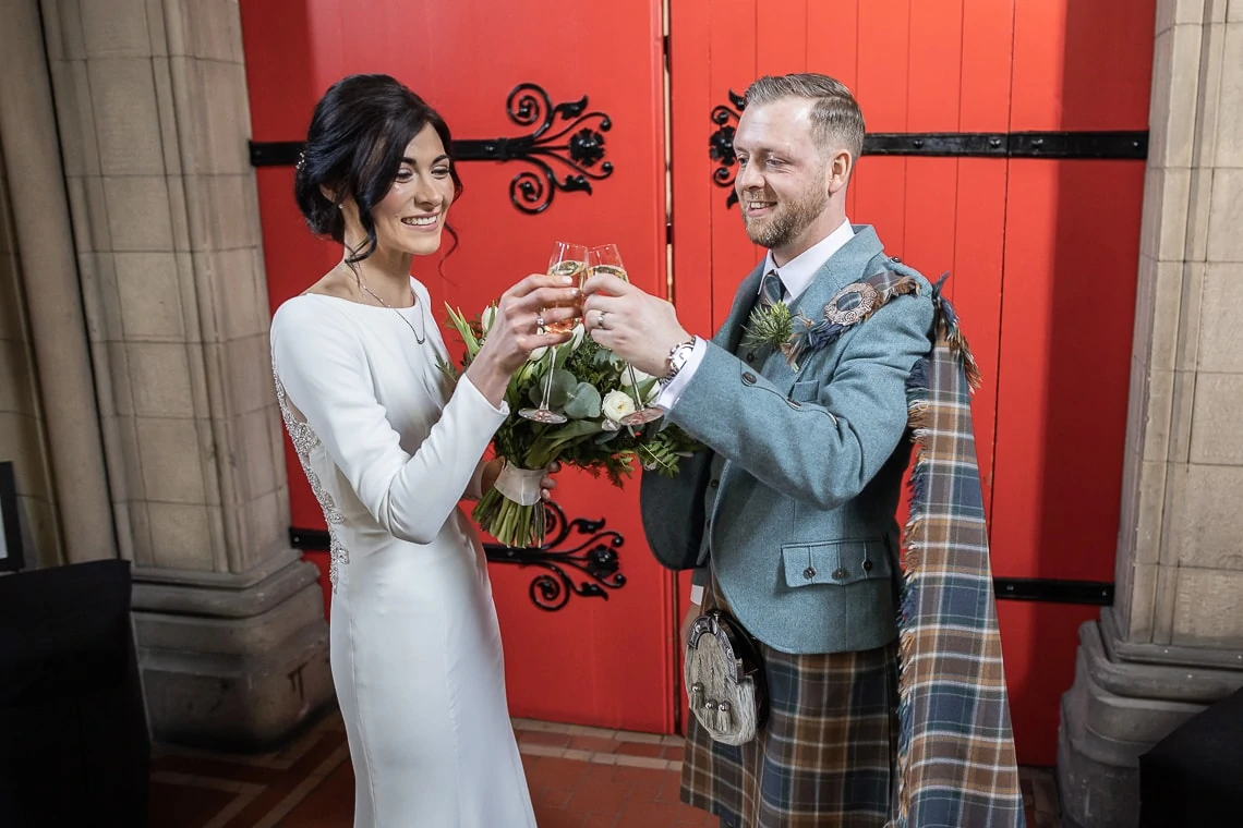 bride and groom raise a toast to each other in front of the red doors at Mansfield Traquair