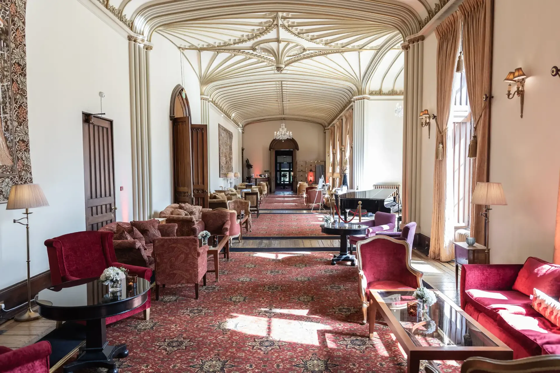 A long, elegant room with arched ceilings, red carpets, and plush seating. Large windows line one side, and ornate chandeliers hang from the ceiling.