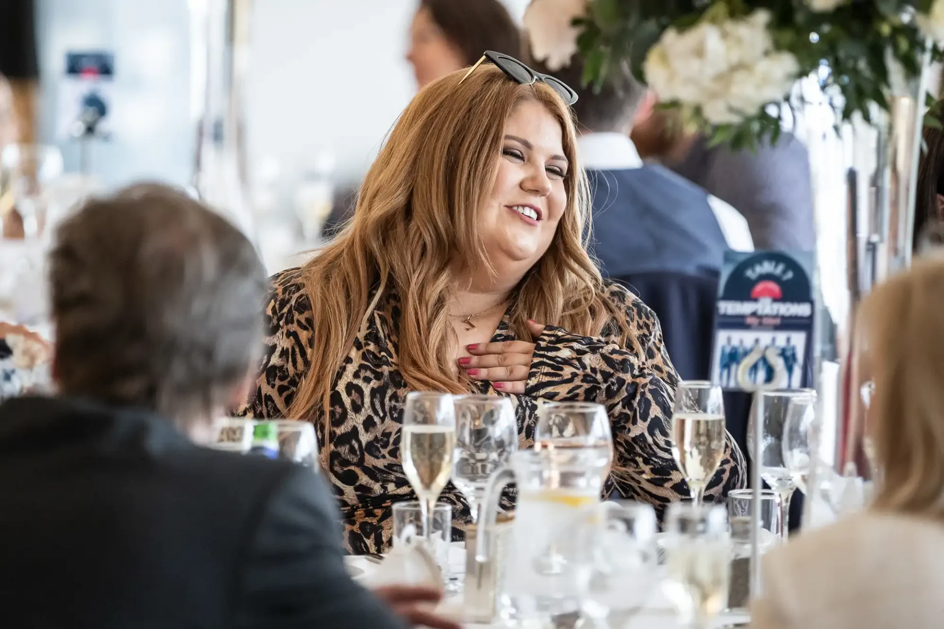 Person with long hair and sunglasses on head, wearing a leopard print outfit, smiling at a table with drinks and flowers during a social event.